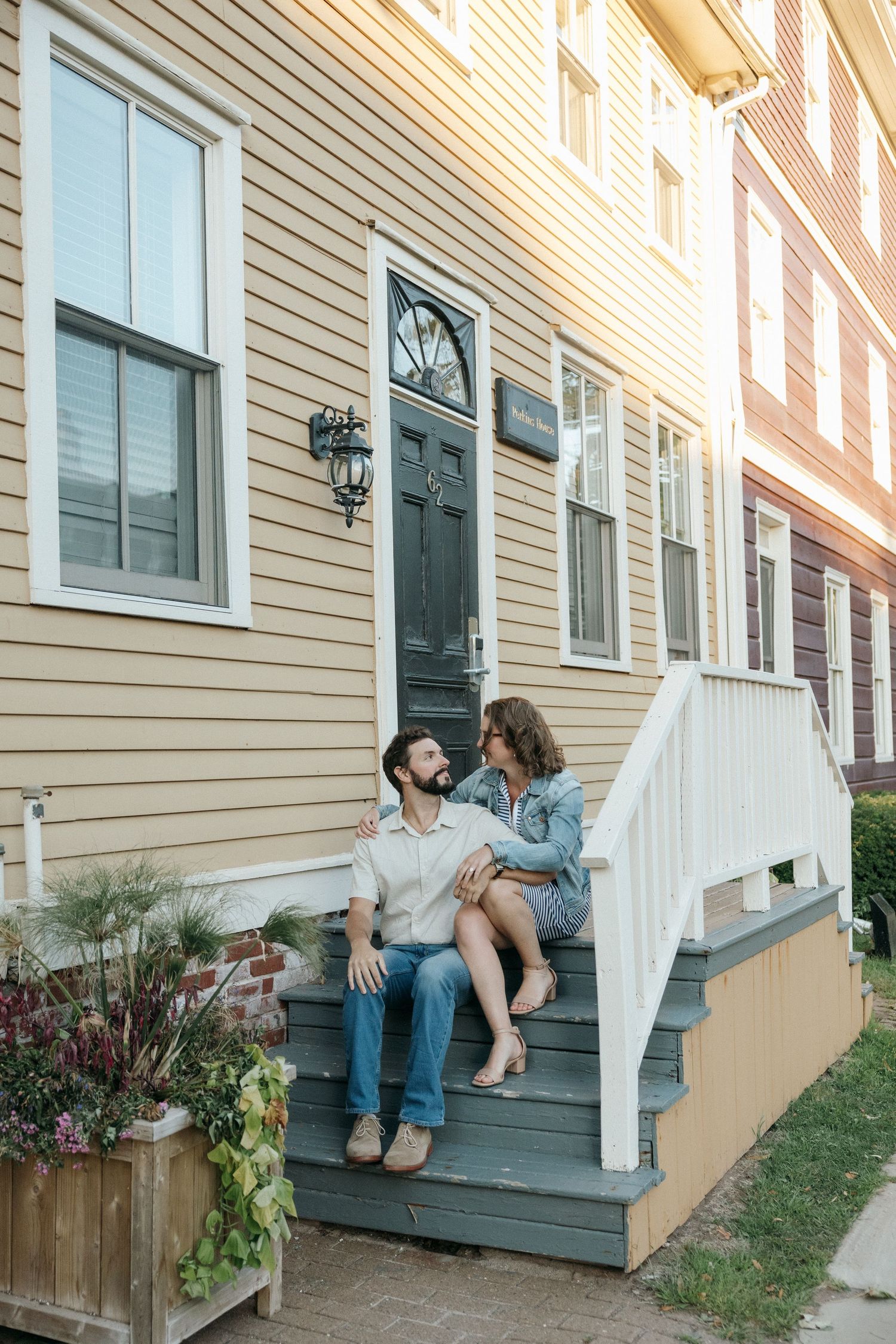 A couple sits together on wooden porch steps of a yellow house with a black door.