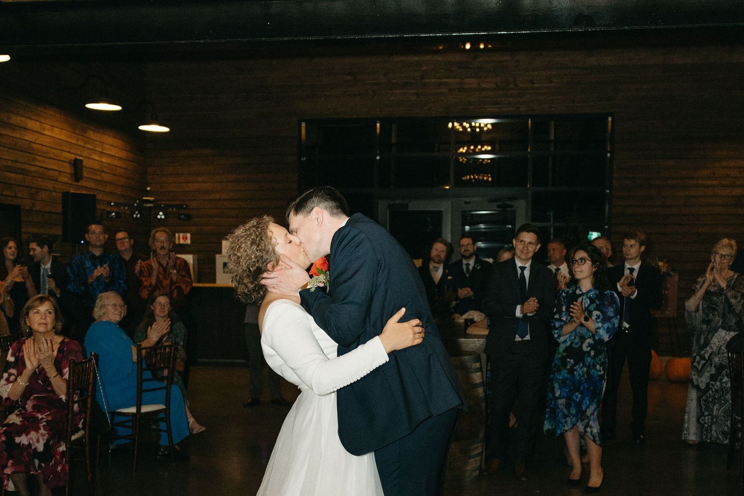Newlyweds dance surrounded by guests in a dimly lit reception venue.