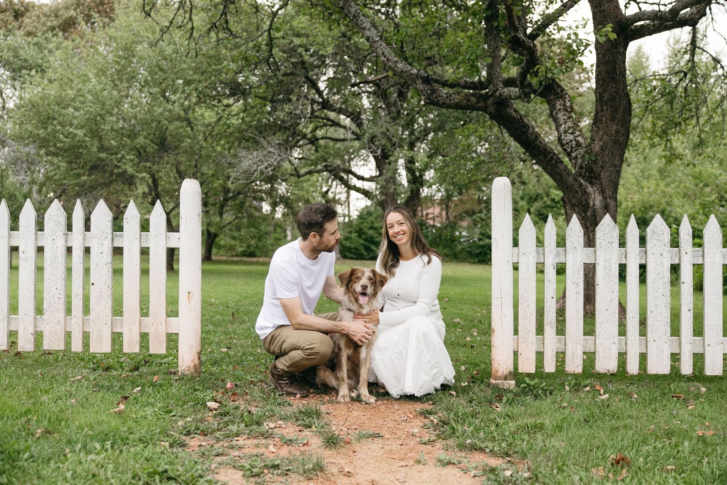 A smiling couple crouches with their dog by a white picket fence surrounded by lush green trees.