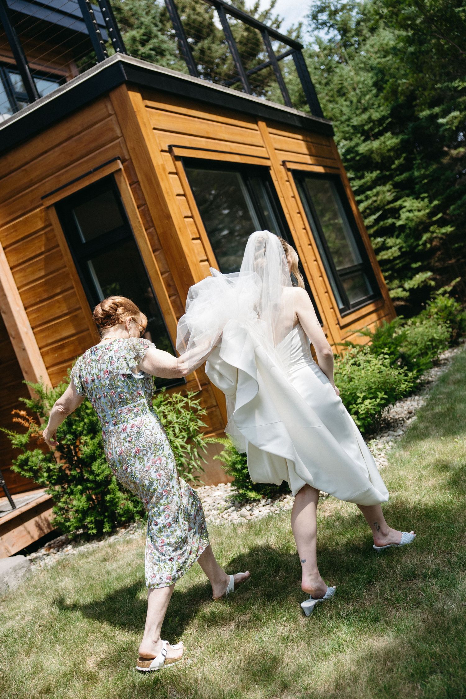 Bride in white gown and veil walks with woman in floral dress toward modern wooden cabin on sunny day.