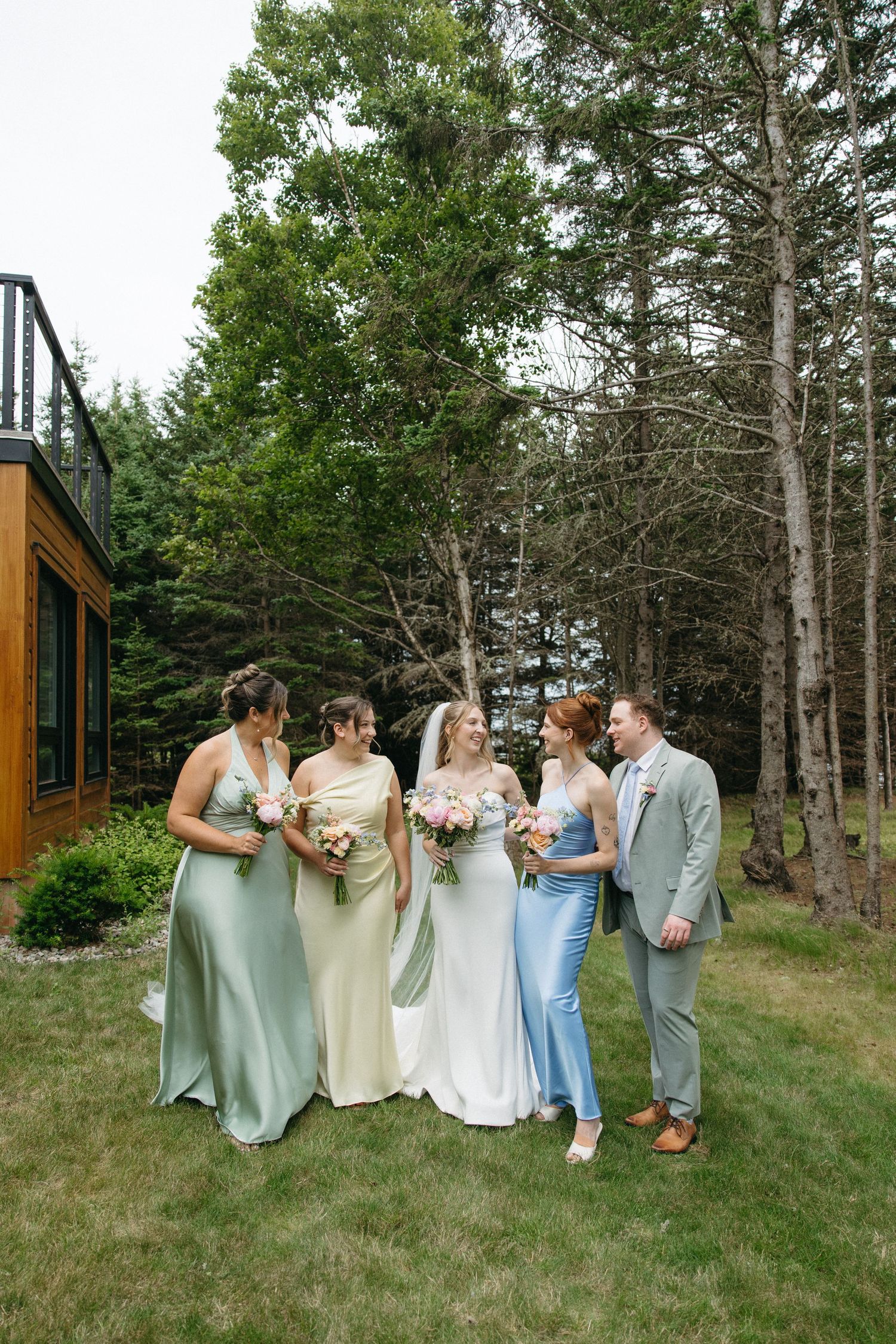 Bride with bridesmaids in sage green and blue gowns holding bouquets outdoors near trees.