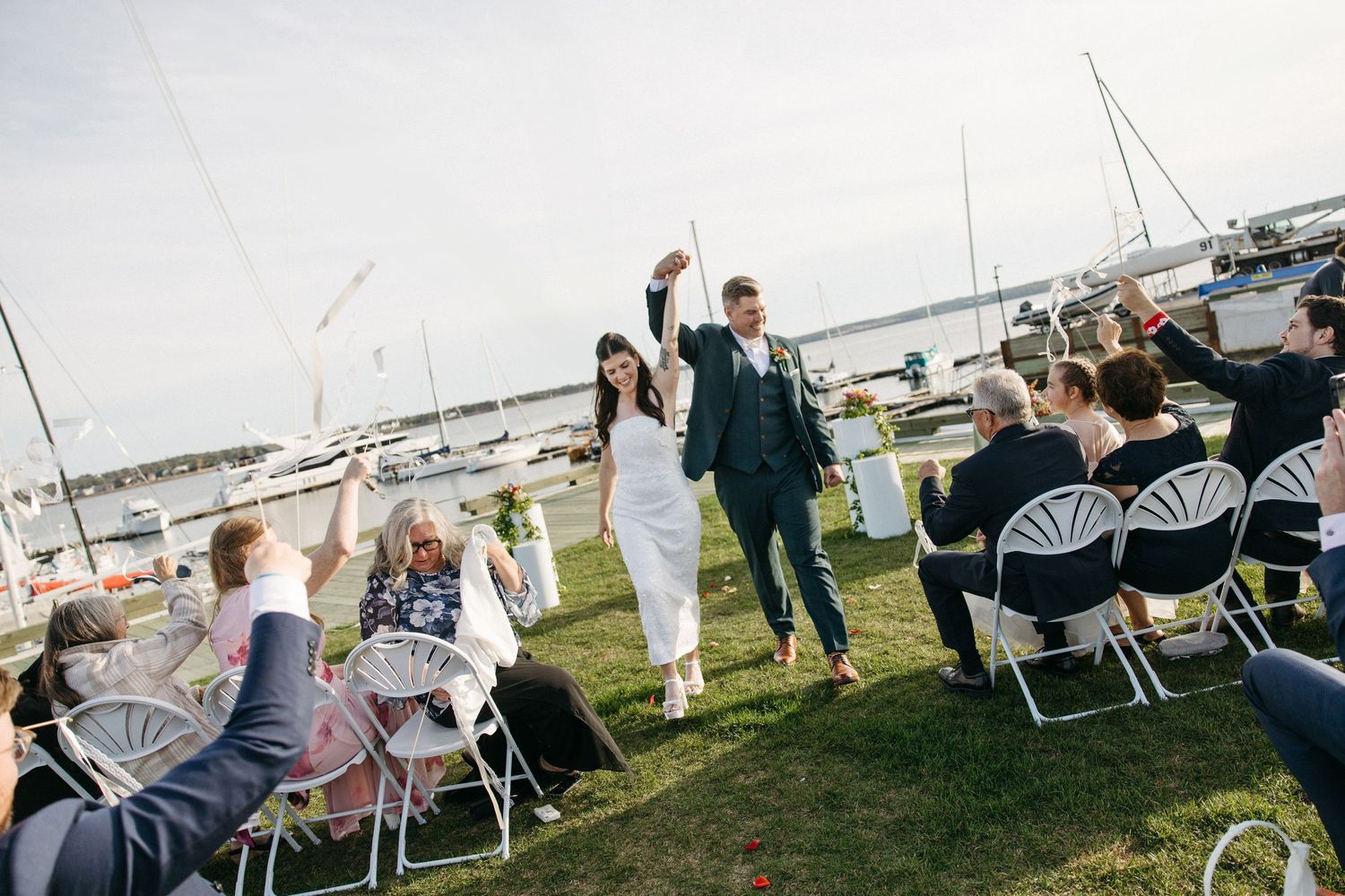 Newlywed couple walks down the grass aisle with raised arms as guests in white chairs cheer at a waterfront wedding with sailboats.