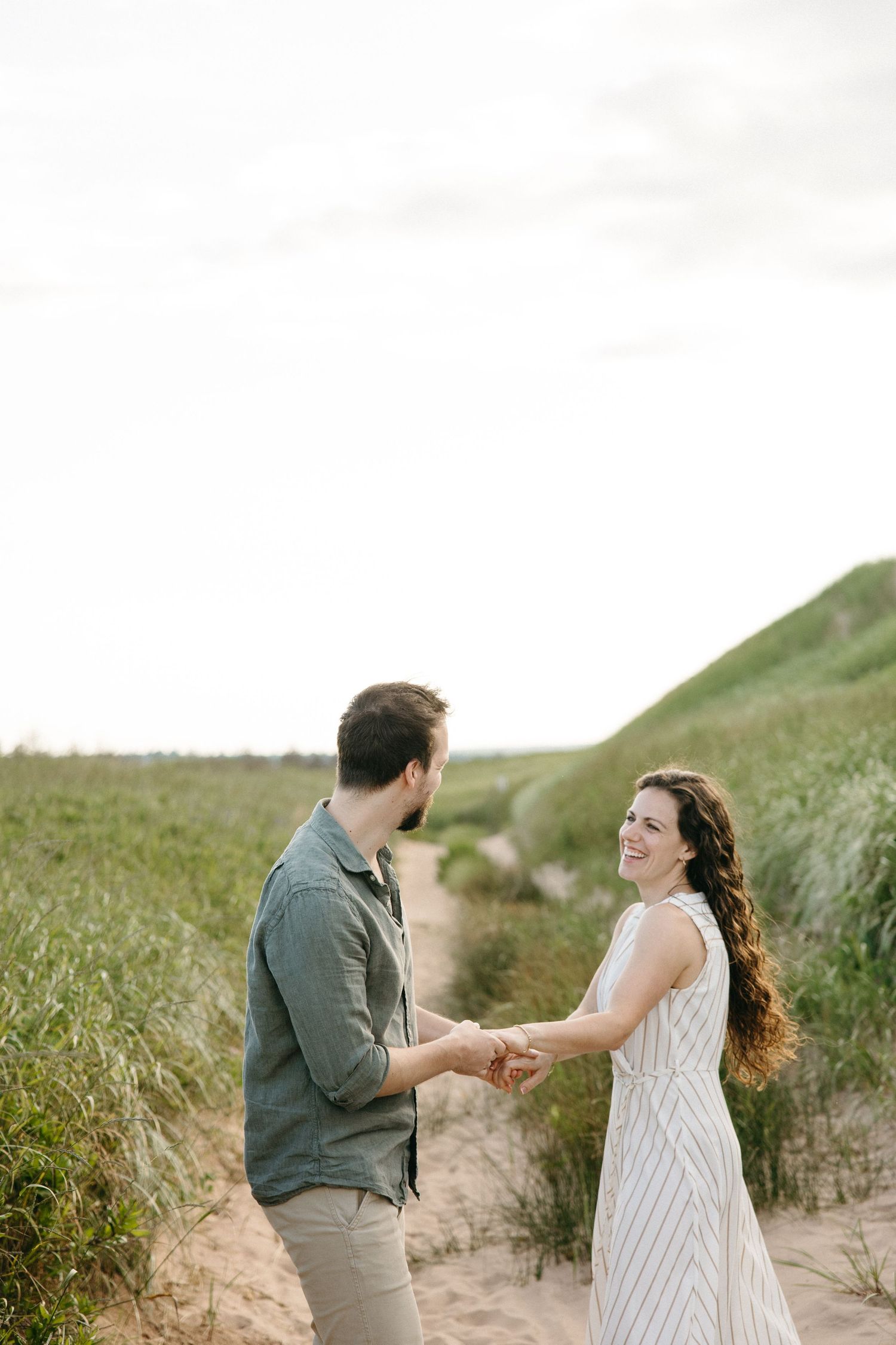 A smiling couple holds hands on a sandy path surrounded by green dunes, woman in striped dress, man in gray shirt.