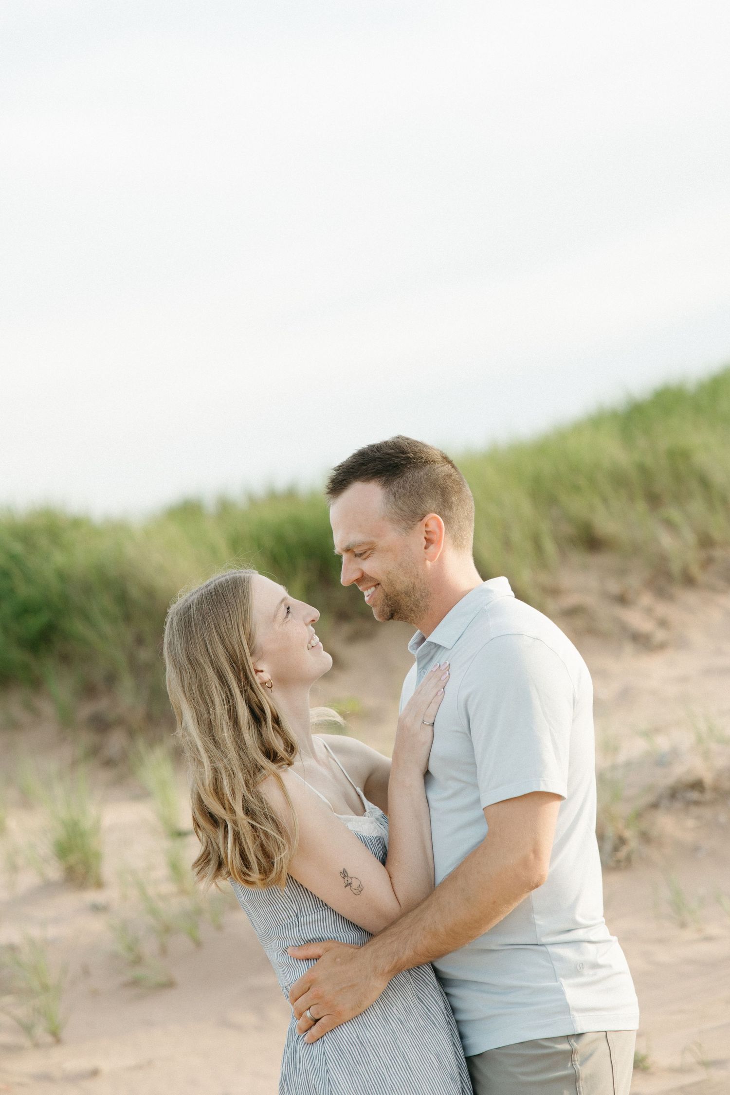 A smiling couple embracing on a sandy beach with grass dunes in the background.