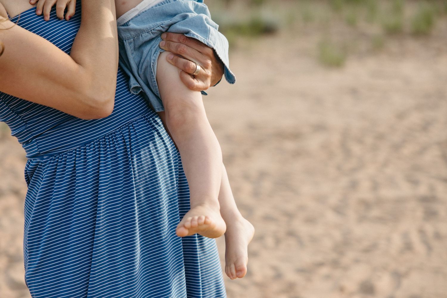 Mother in striped blue dress holding young barefoot child on sandy beach.