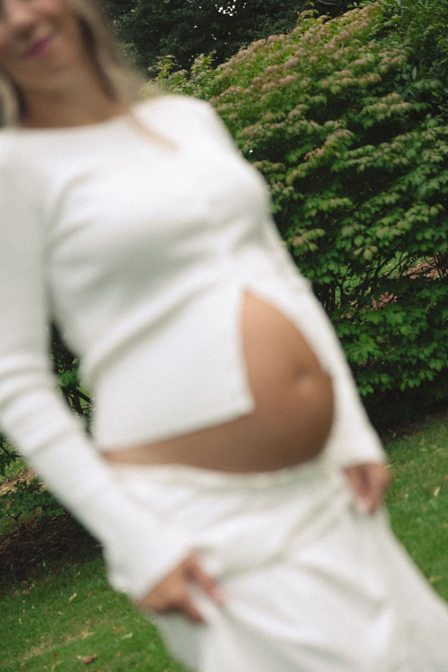 Pregnant woman in white two-piece outfit showing baby bump, standing outdoors near green bushes.