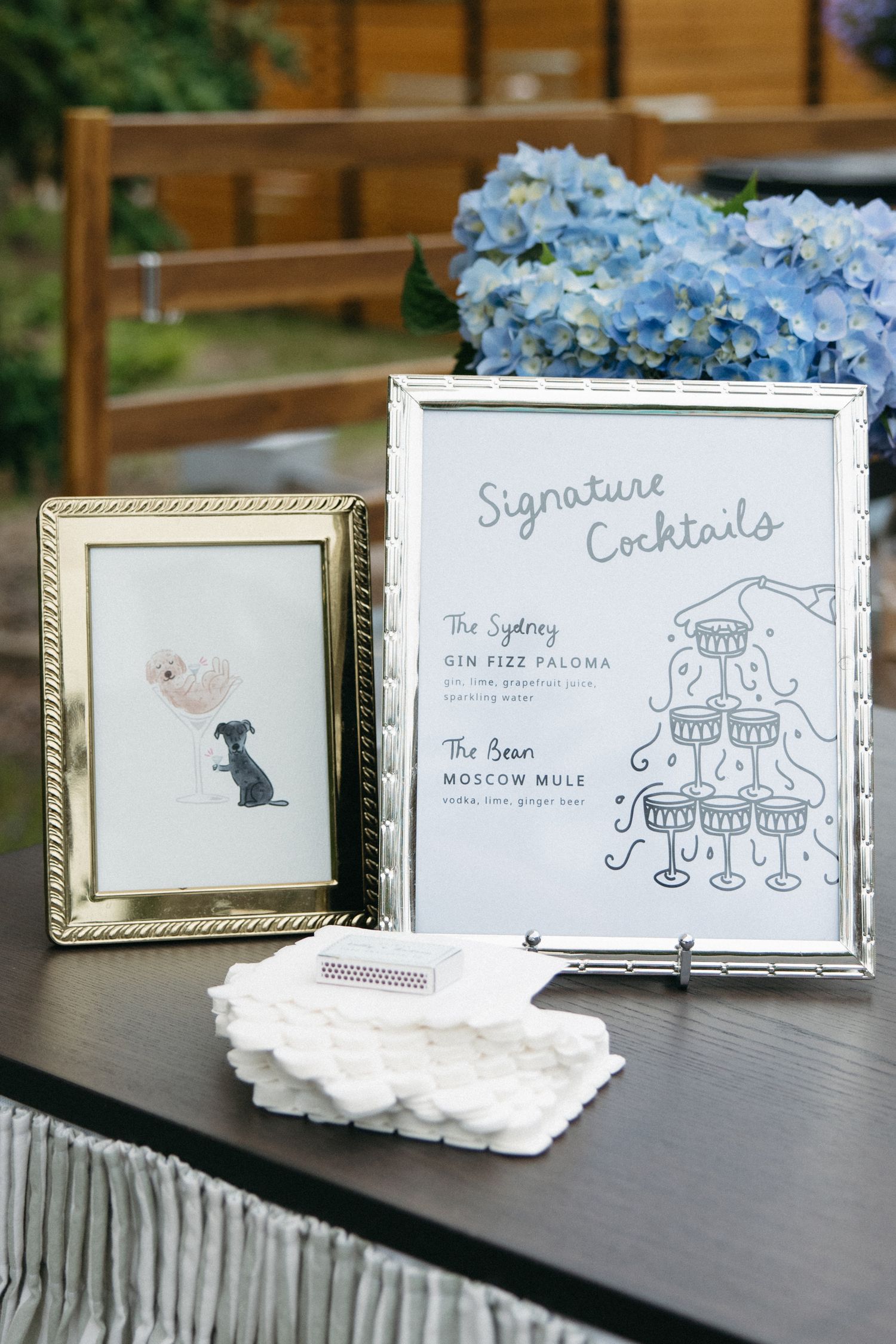 Wedding signature cocktails sign in silver frame beside dog portrait and blue hydrangeas.