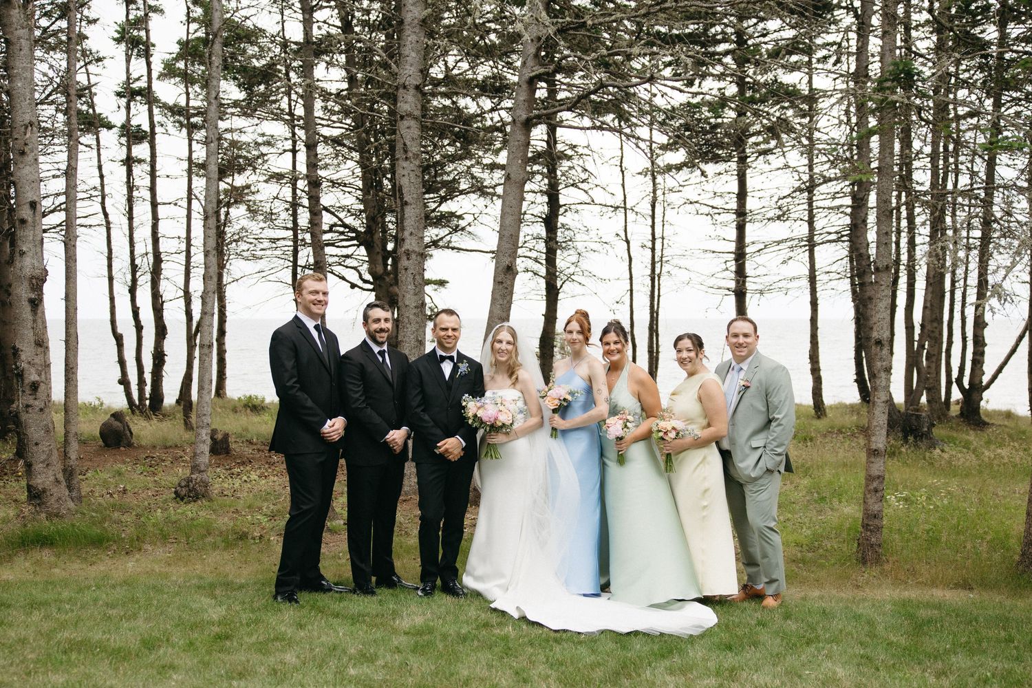Wedding party of seven posing together in formal attire among pine trees with ocean in background.