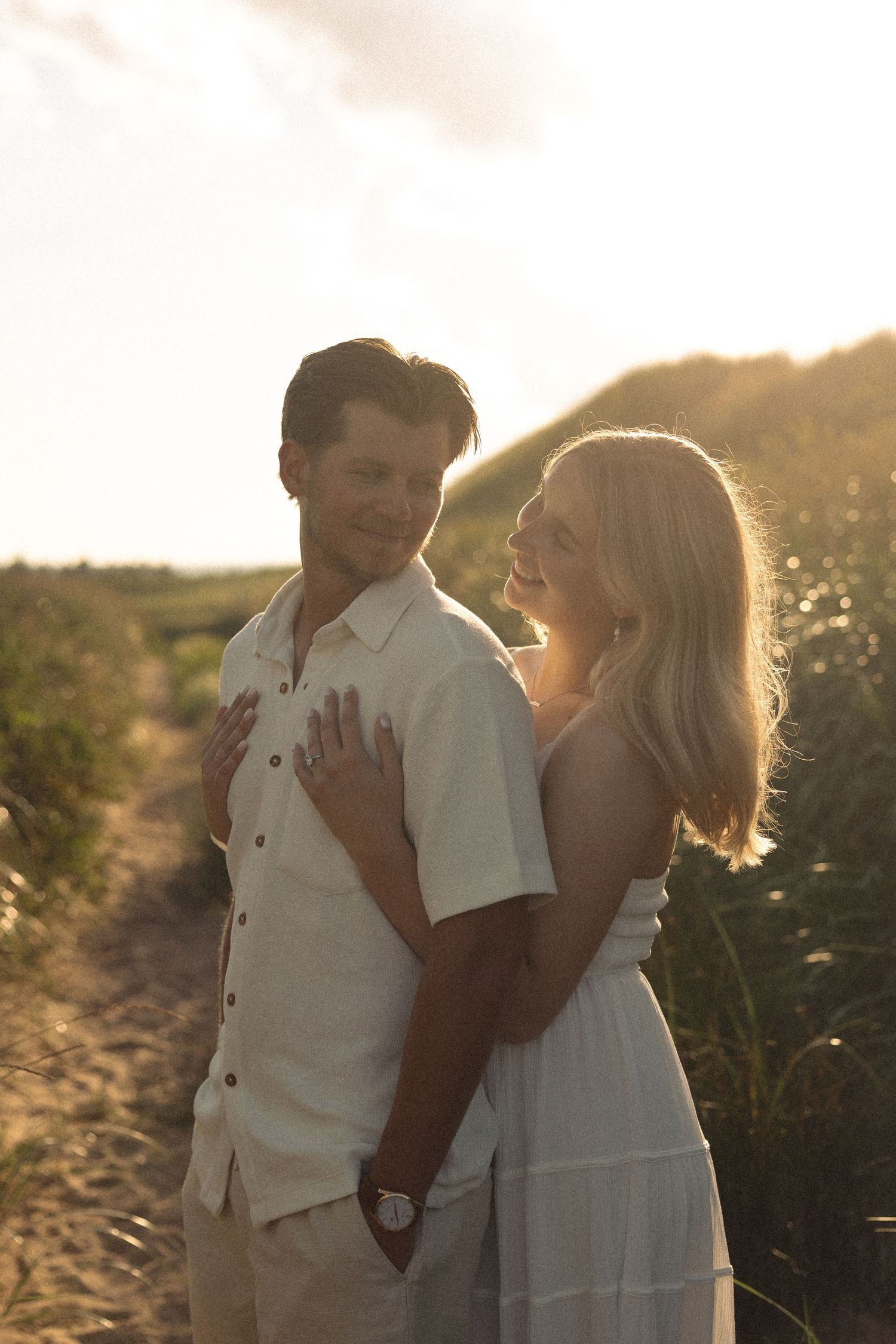 Engaged couple embracing on sandy beach path surrounded by golden sunset light.