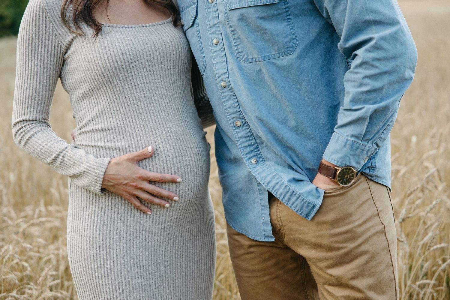 Pregnant couple standing in a golden wheat field, woman in grey ribbed dress cradling baby bump.