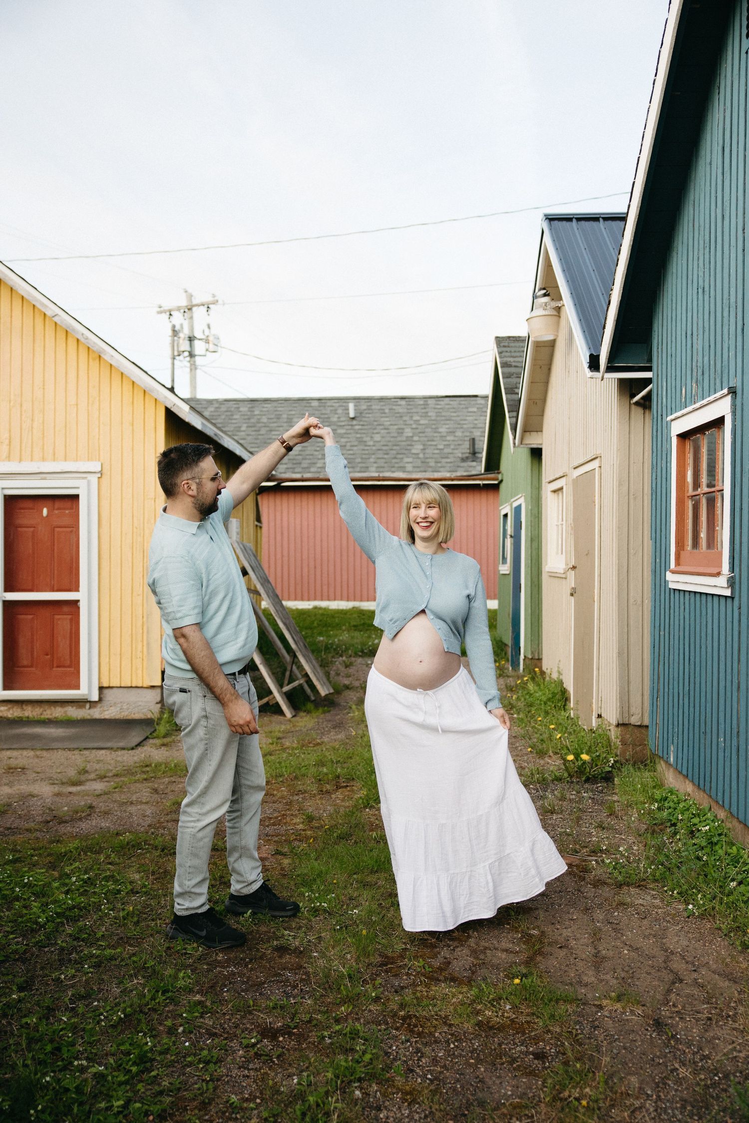 A pregnant woman dances with her partner outdoors among colorful wooden buildings.