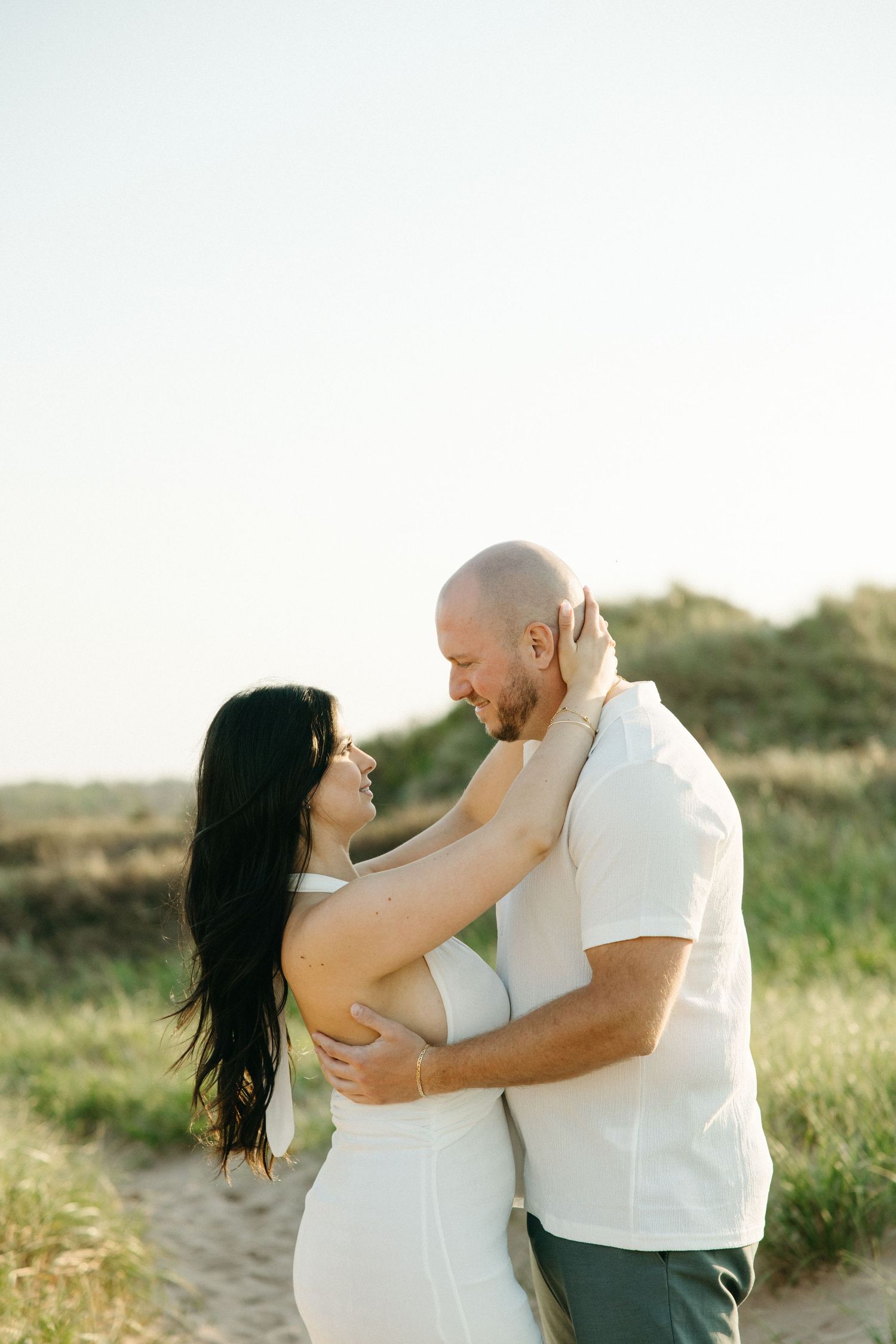 Couple embracing romantically on a sandy beach path at golden hour, woman in white dress with long dark hair.