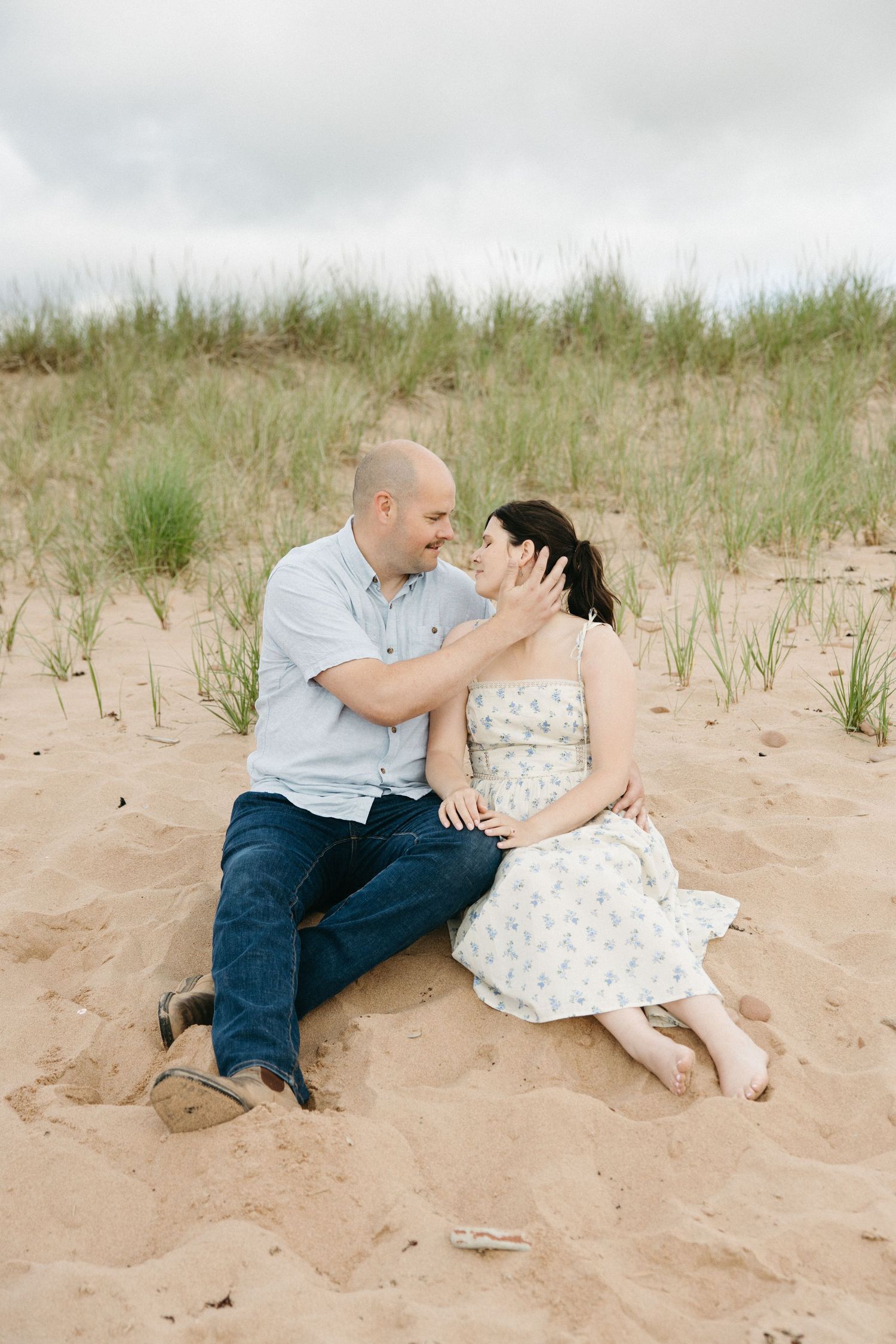 Couple sitting on sandy beach dunes, smiling at each other on a cloudy day.