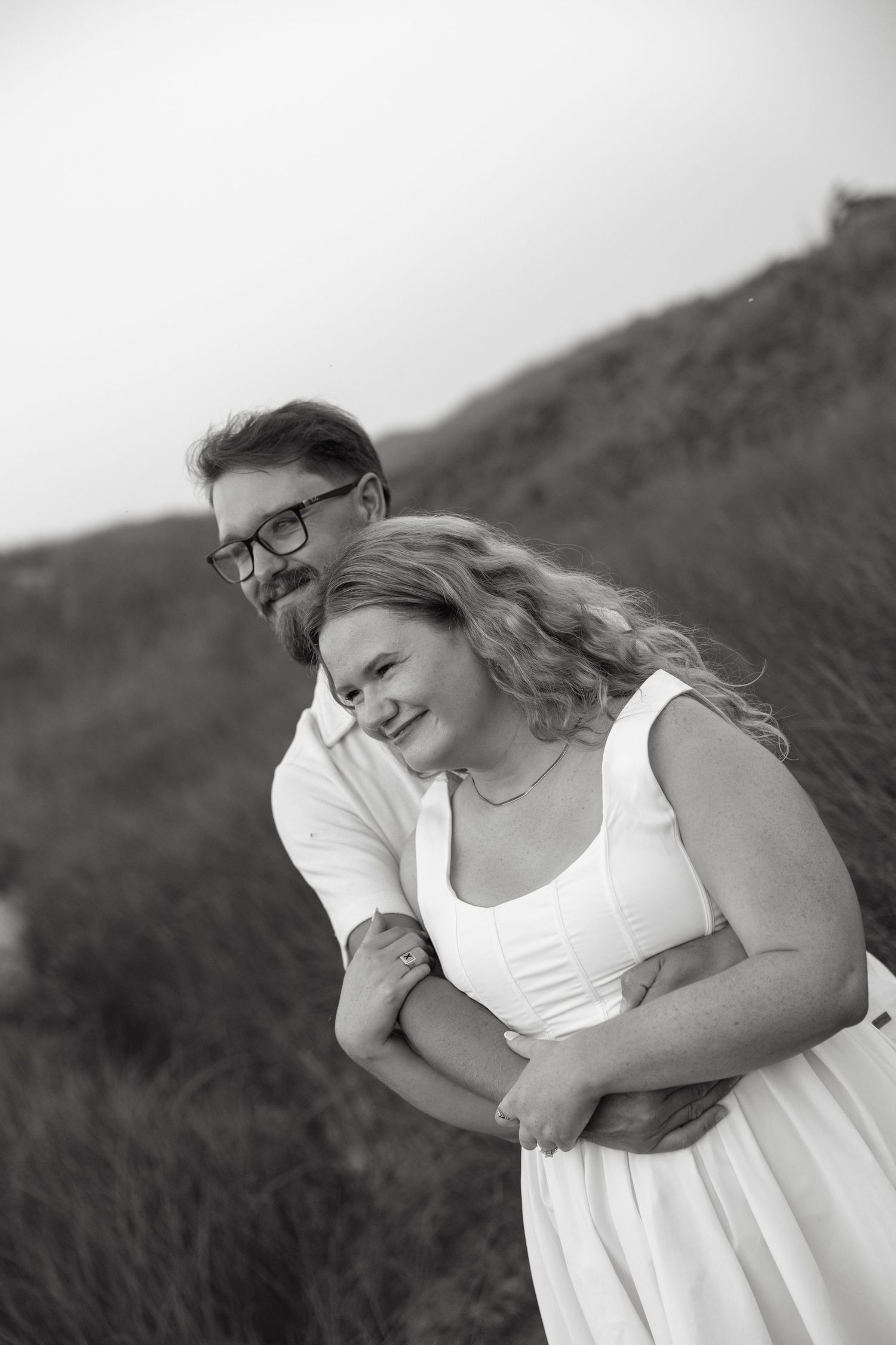 Black and white photo of smiling couple embracing in grassy dunes, man hugging woman from behind.