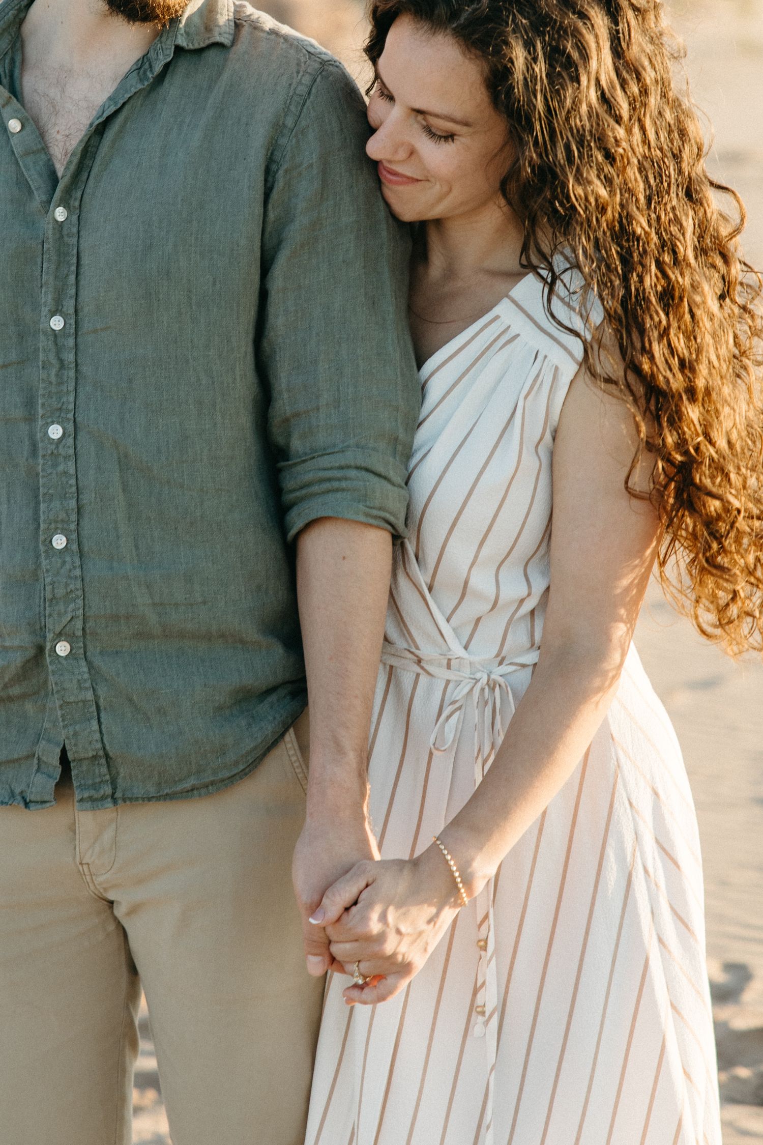 Couple holding hands at beach, woman in striped dress leans on mans shoulder in golden sunlight.