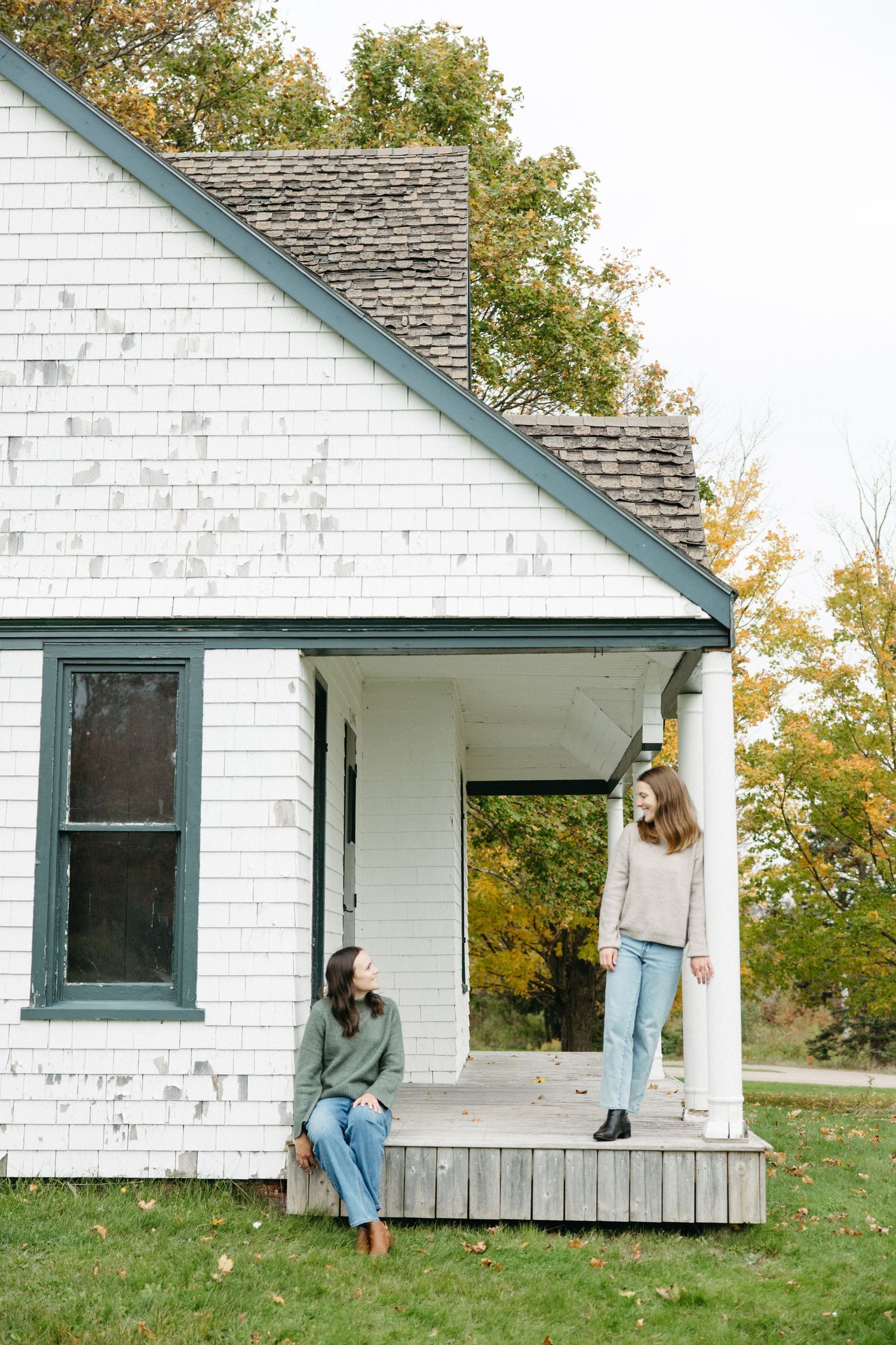 Two women in sweaters and jeans relax on the porch of a white clapboard house with green trim, surrounded by autumn trees.