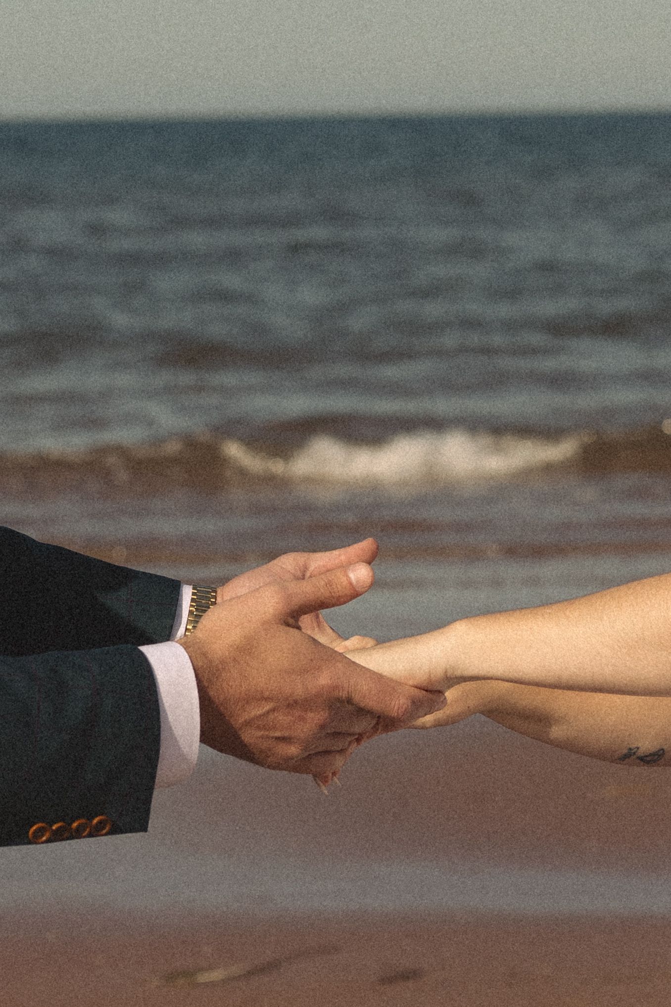 Close-up of a groom in a navy suit gently holding a partner's hand on a sandy beach with ocean waves in the background.
