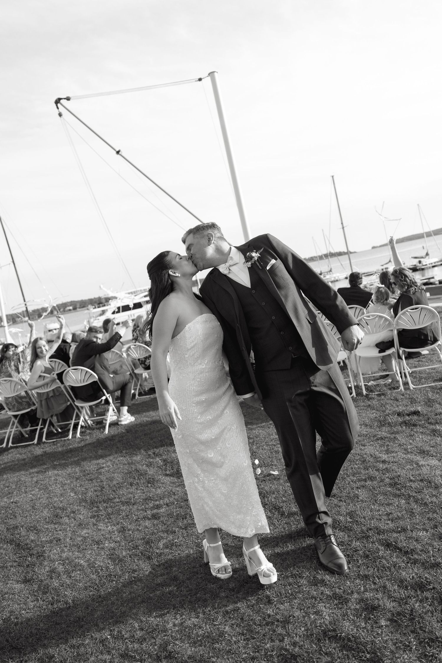 Black and white photo of a bride and groom kissing while walking past guests seated in chairs at an outdoor waterfront wedding ceremony.