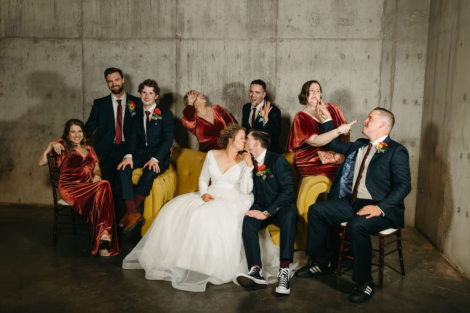 Wedding party poses casually on yellow couch against concrete wall in modern venue.