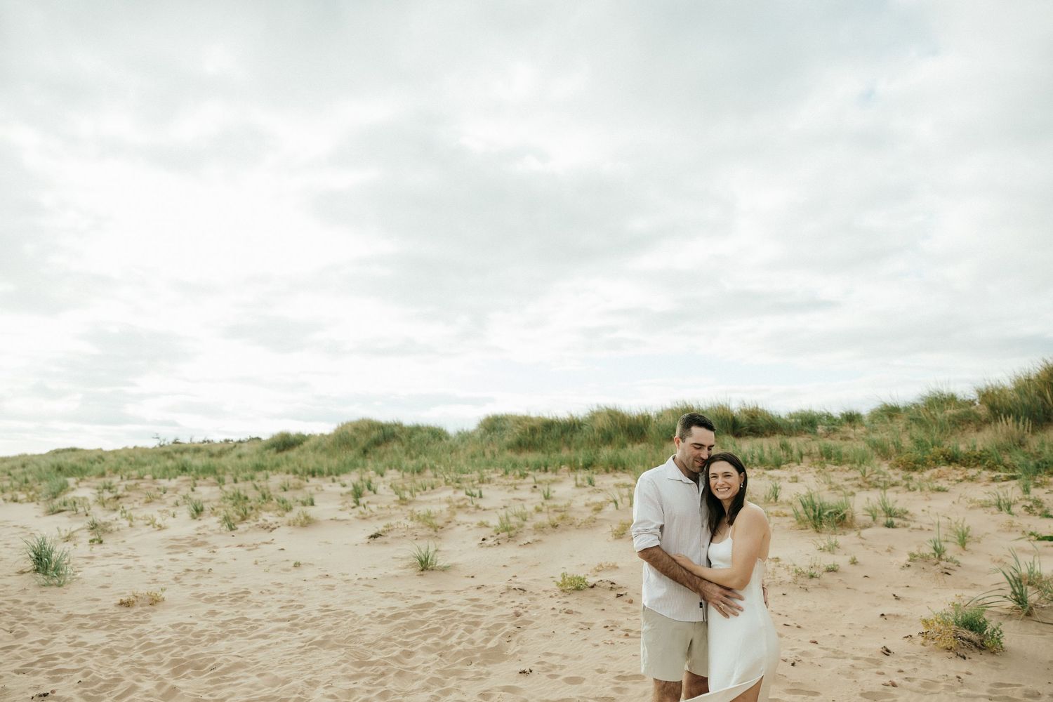 A romantic couple embraces on a sandy beach with grass-covered dunes in the background.
