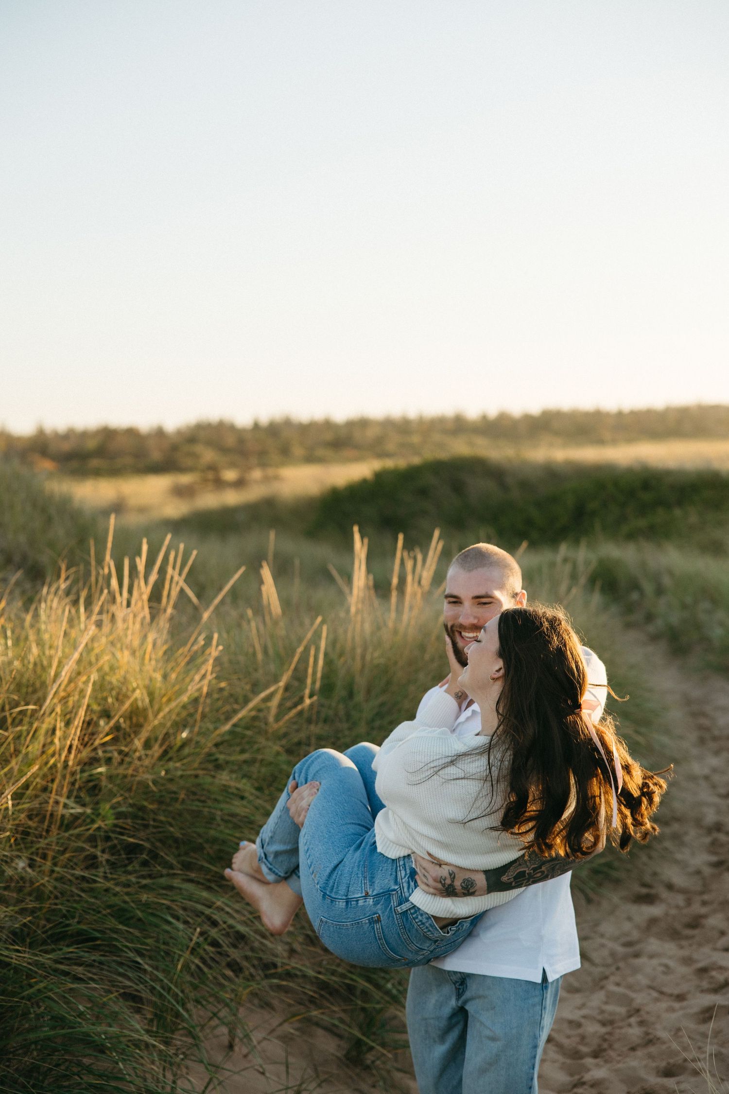 Couple embraces playfully in golden beach grass during sunset photoshoot.