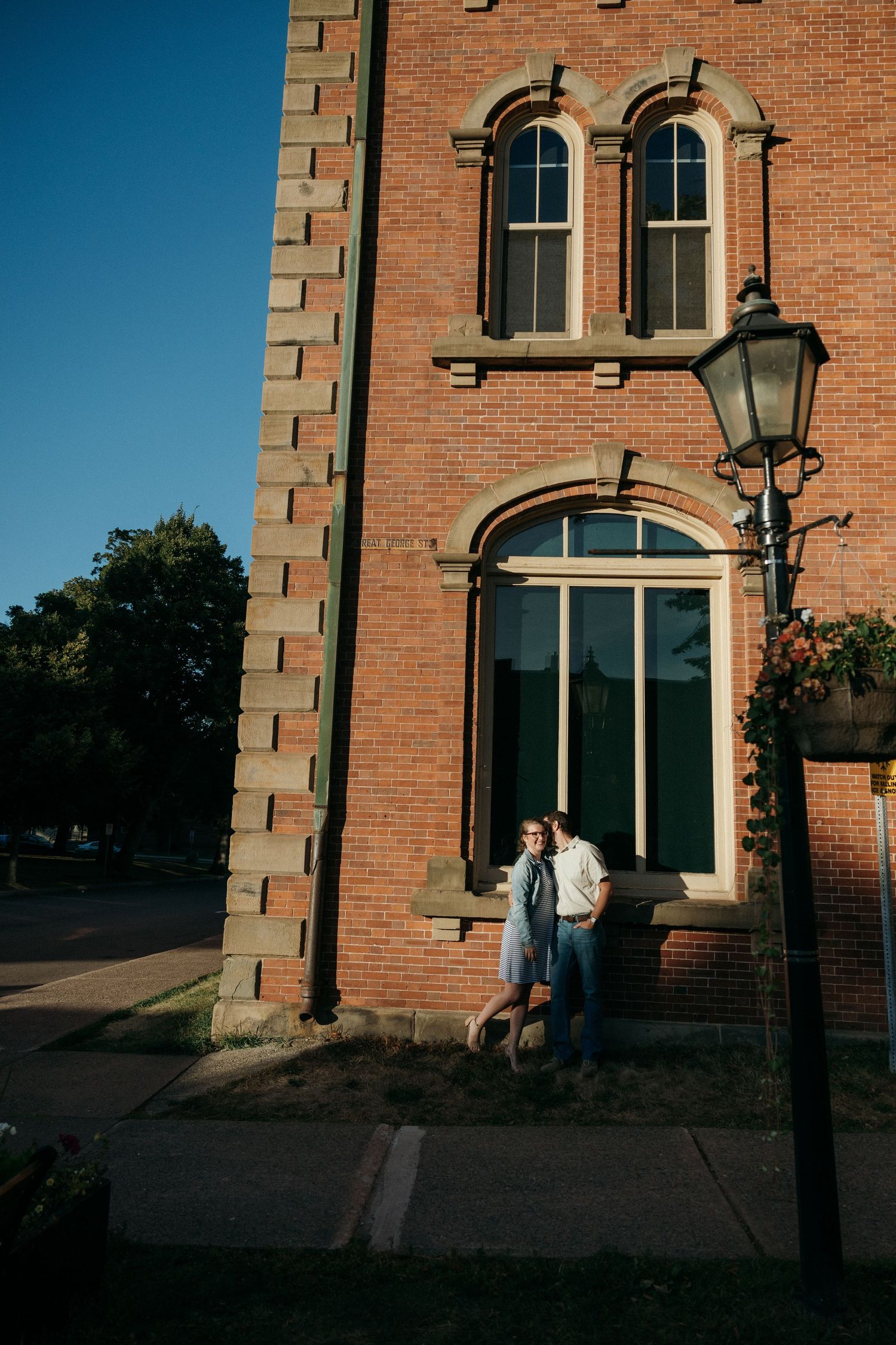 A couple poses against a historic brick building with arched windows at sunset.