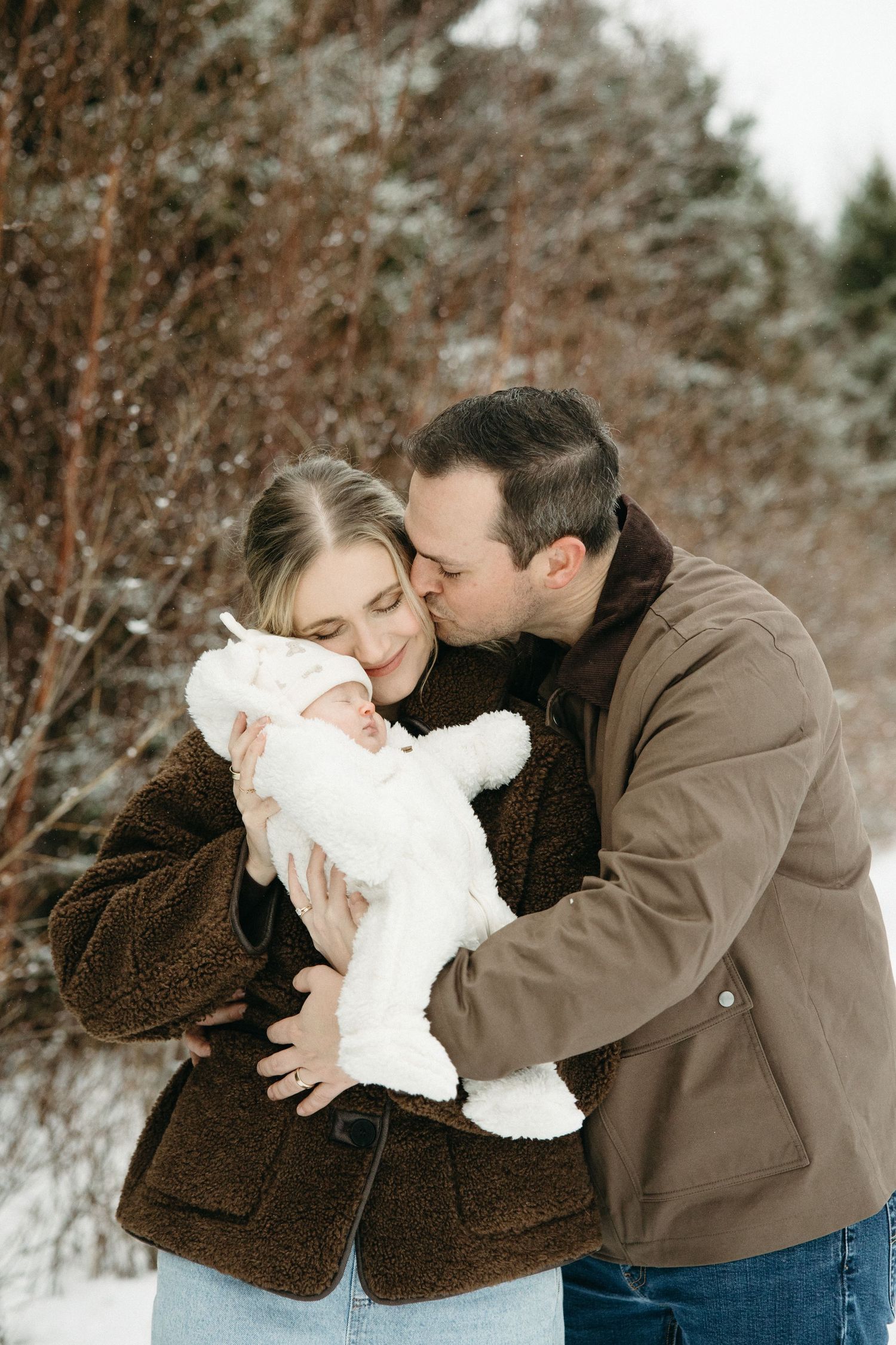 Intimate family moment captured in snowy setting with baby in white winter clothing.