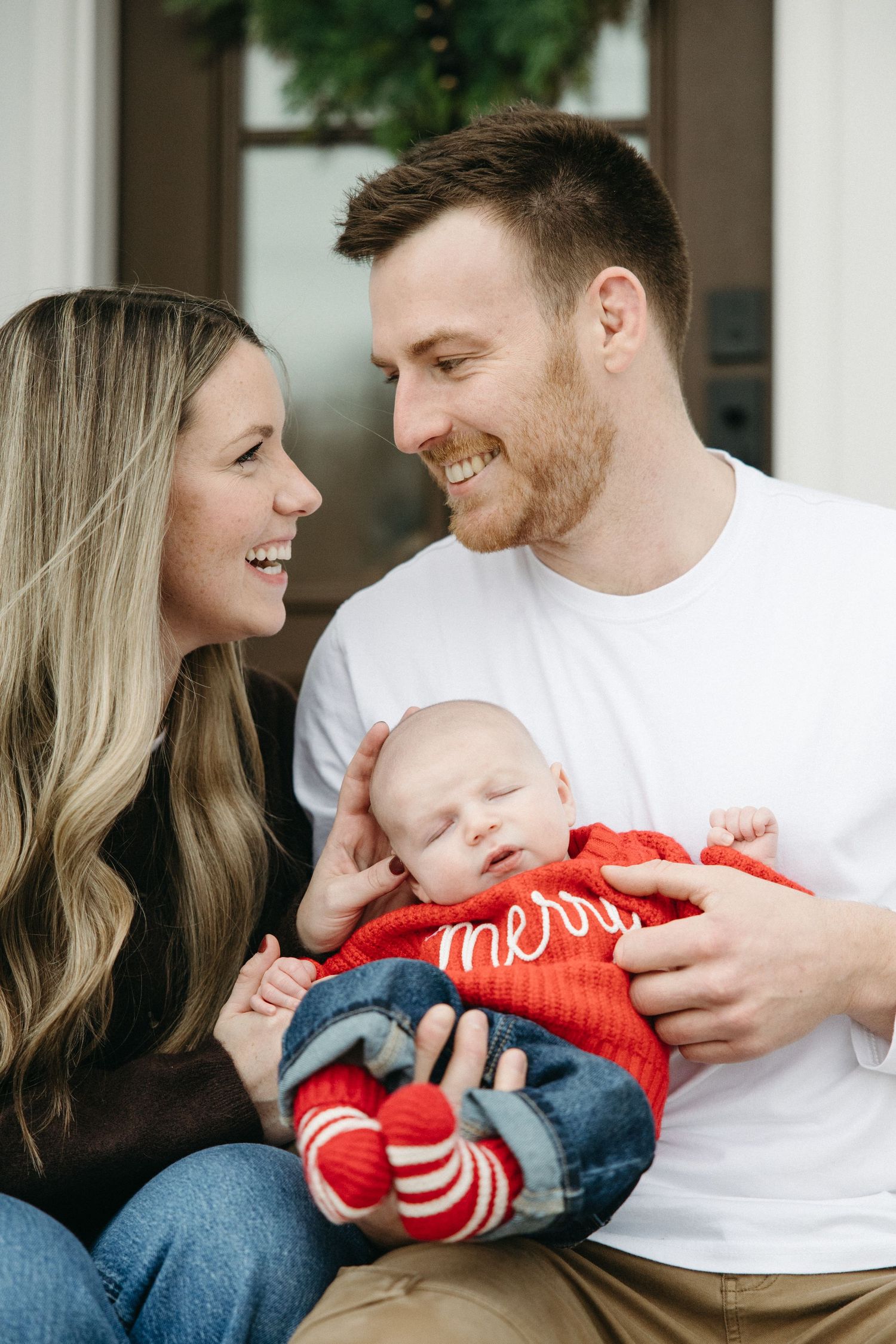 A couple shares tender moments with a baby wearing a red sweater on a porch.