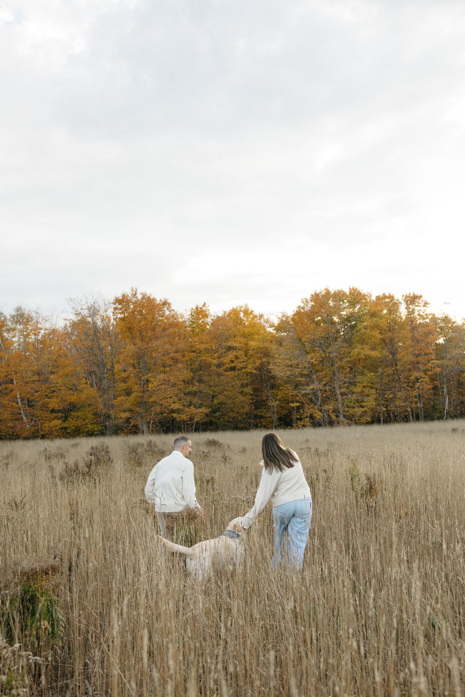 Couple and their dog walking through tall grass meadow with autumn trees in background.