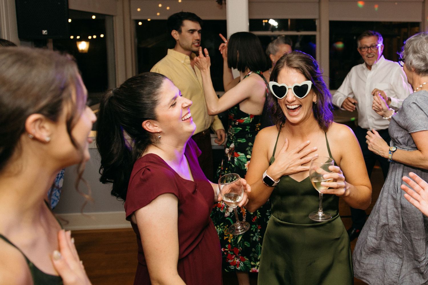 Guests laugh and dance at a wedding reception, one wearing white heart-shaped sunglasses holding champagne.