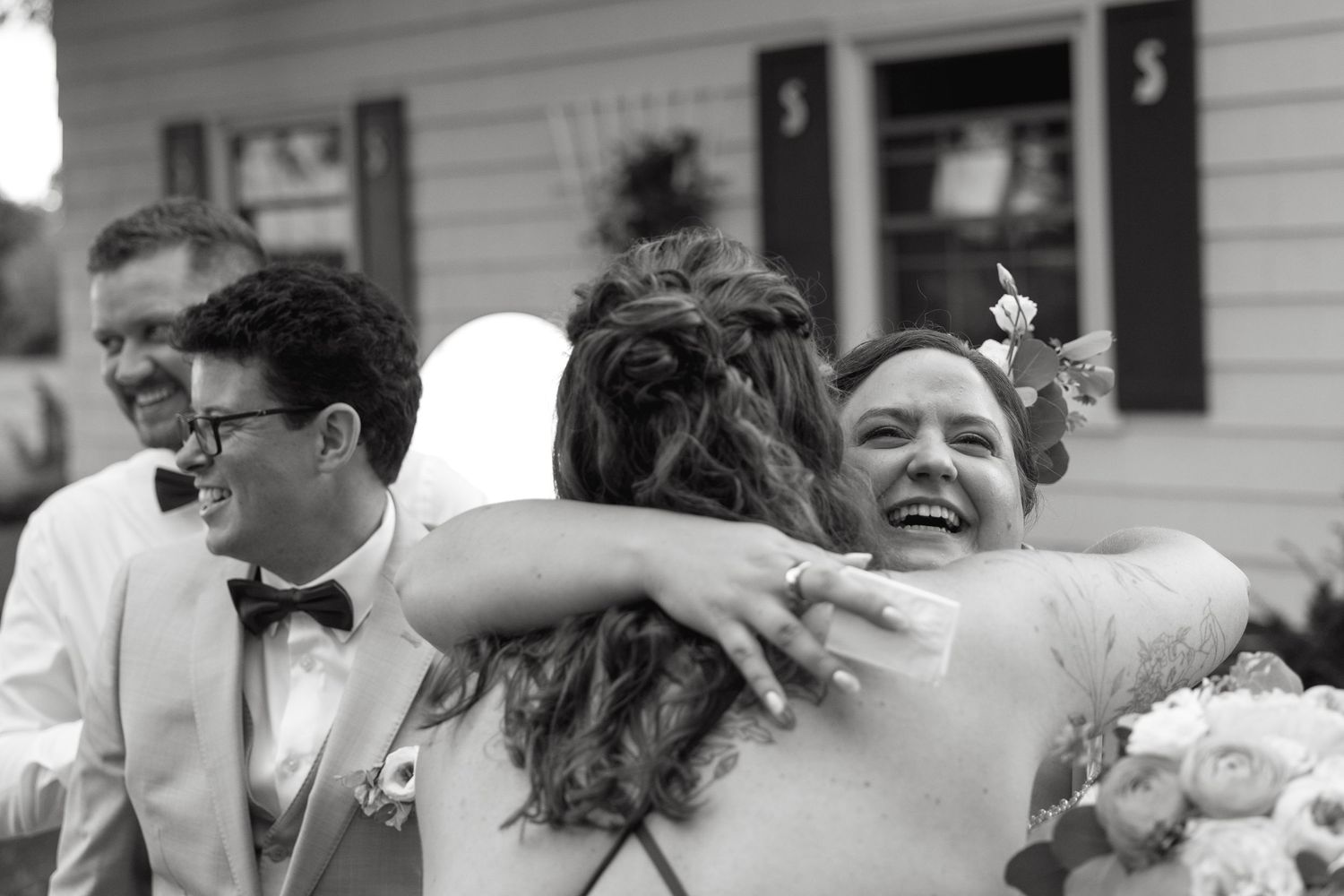 Joyful wedding guest hugging the bride while groom smiles nearby in black and white photo.