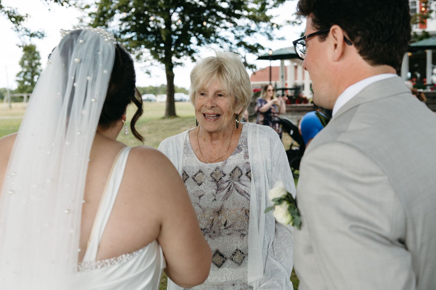 Elderly woman officiant smiling at bride and groom during outdoor wedding ceremony.