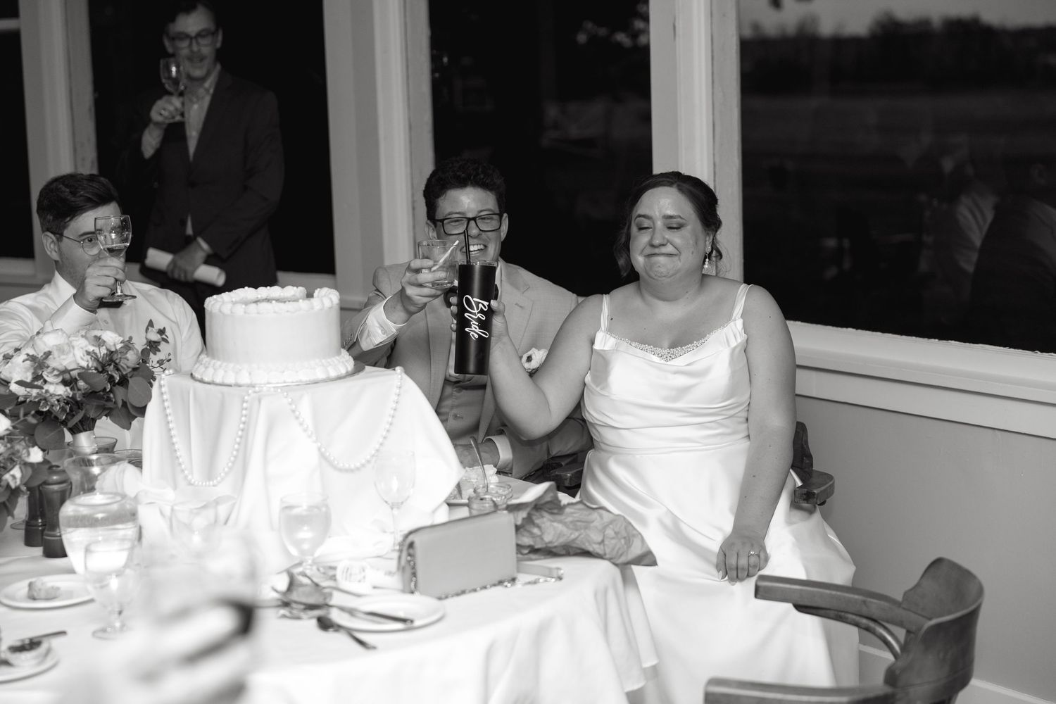 Bride and groom raising glasses during wedding reception toast beside white tiered cake, black and white photo.
