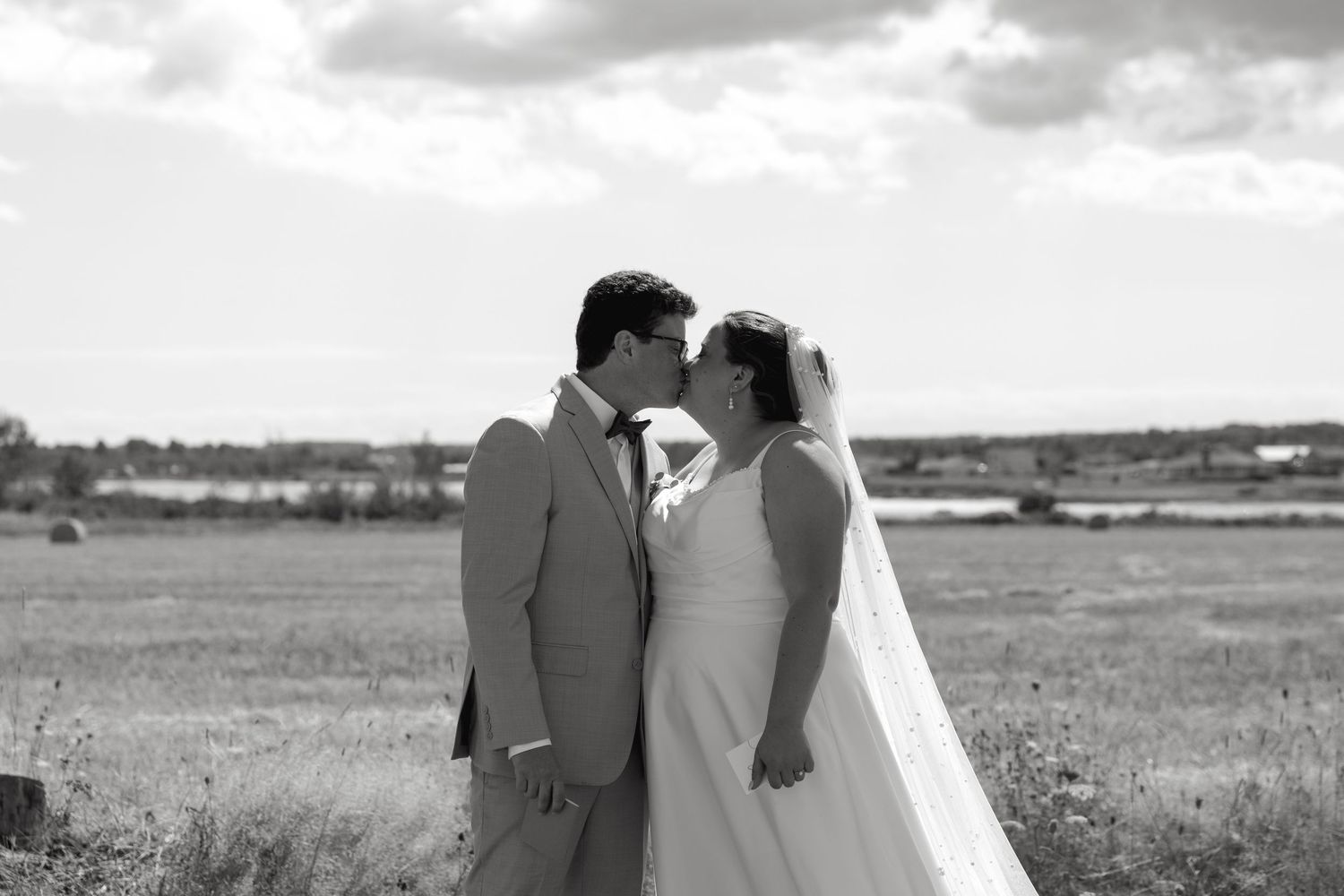 Black and white romantic photo of bride and groom nearly kissing in a sunny golden field with coastal backdrop.