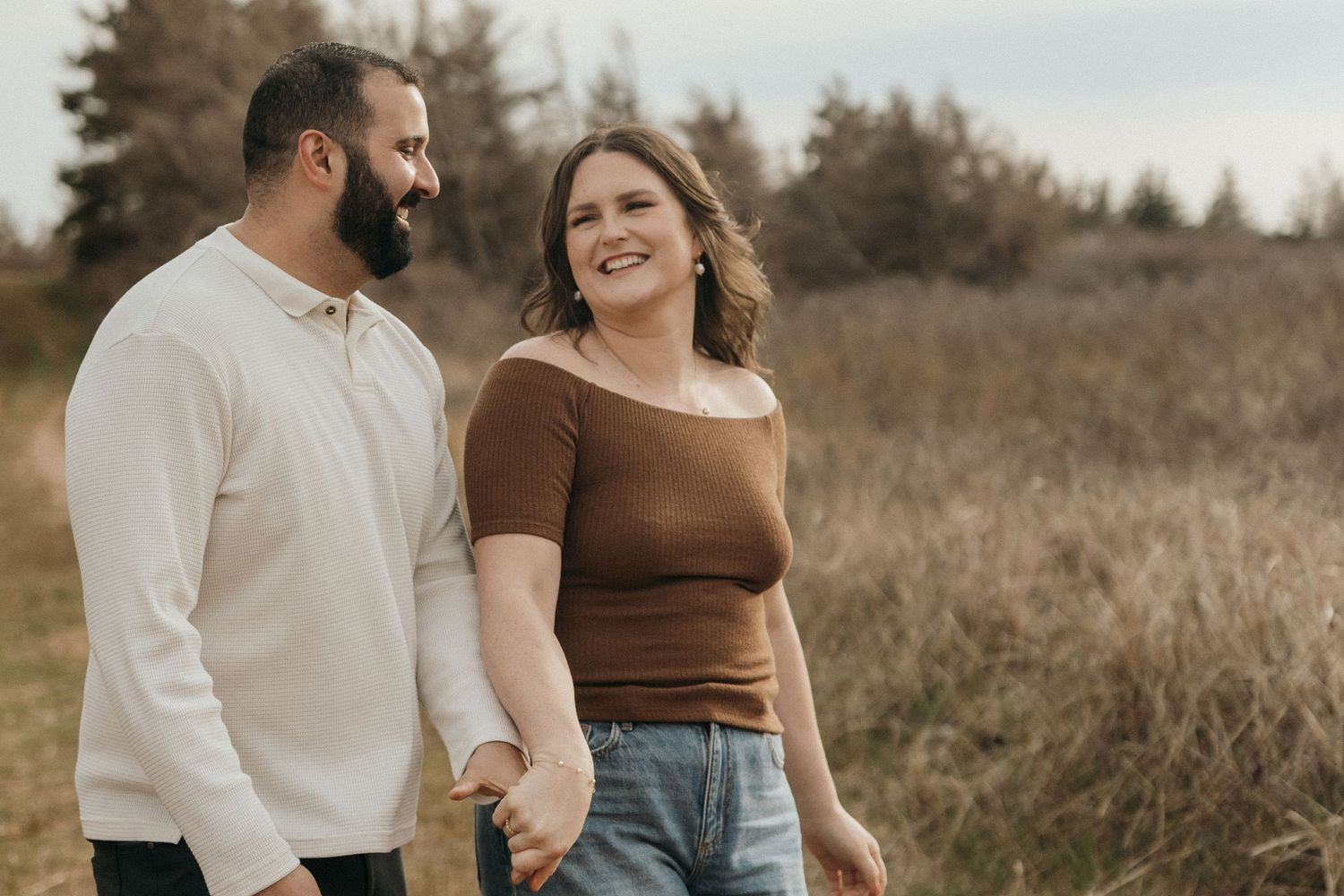 A couple laughs together in a field of tall grass on an overcast day.