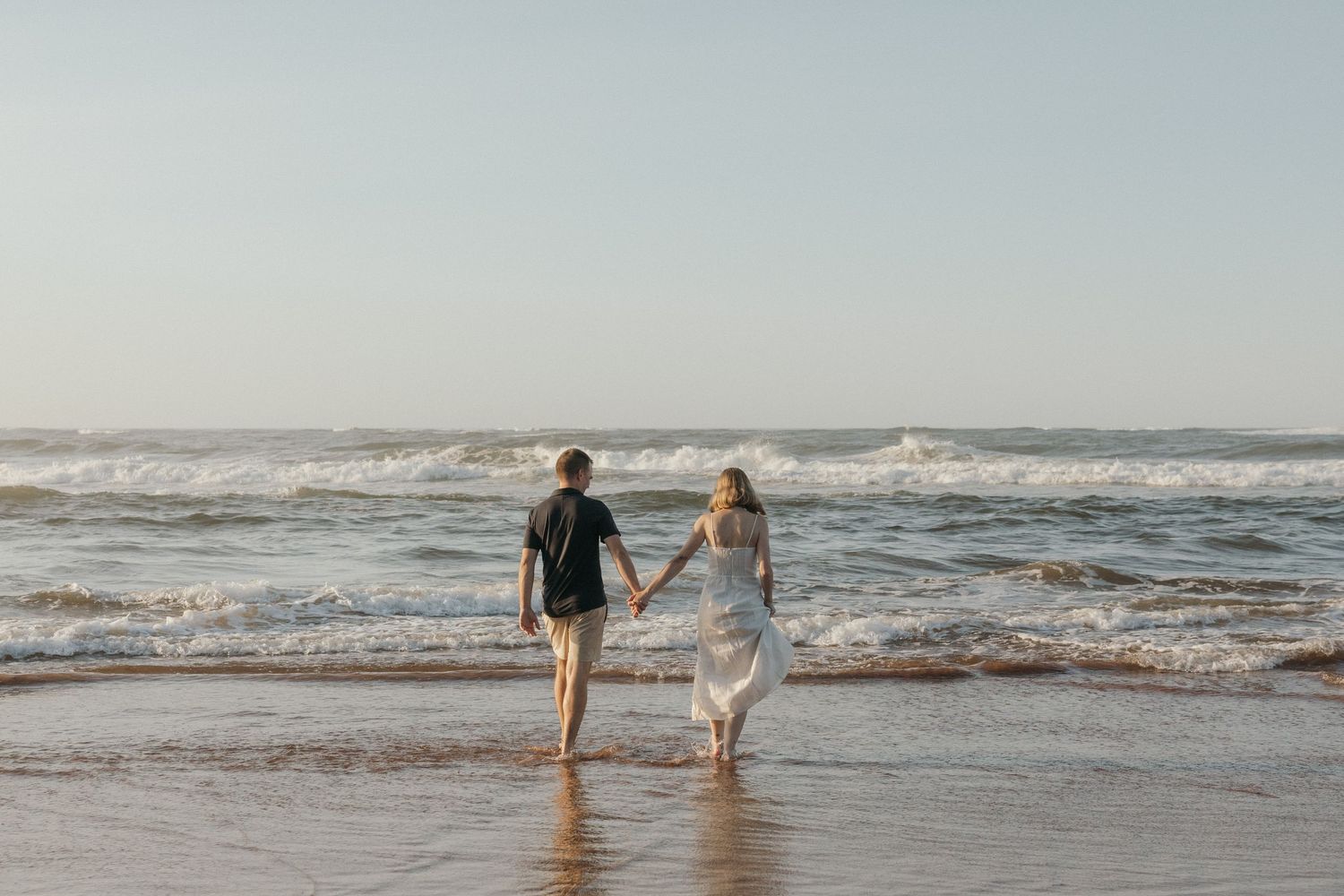 Two figures walk together along a sandy beach with gentle waves rolling in at sunset.