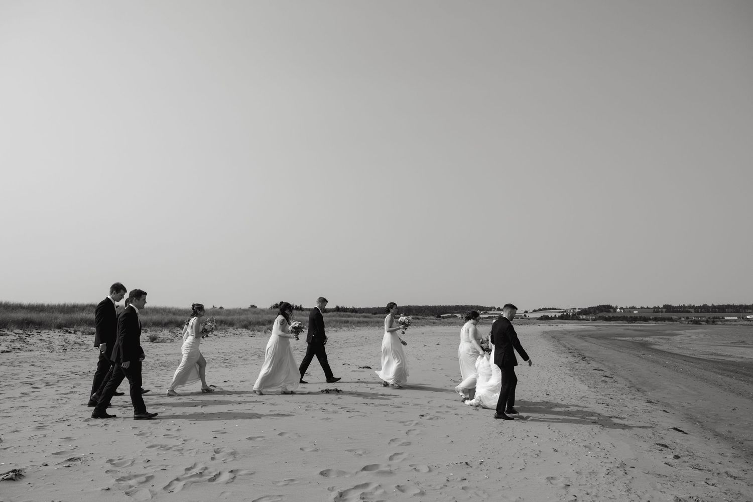 Black and white photo of wedding party walking in line along a beach.