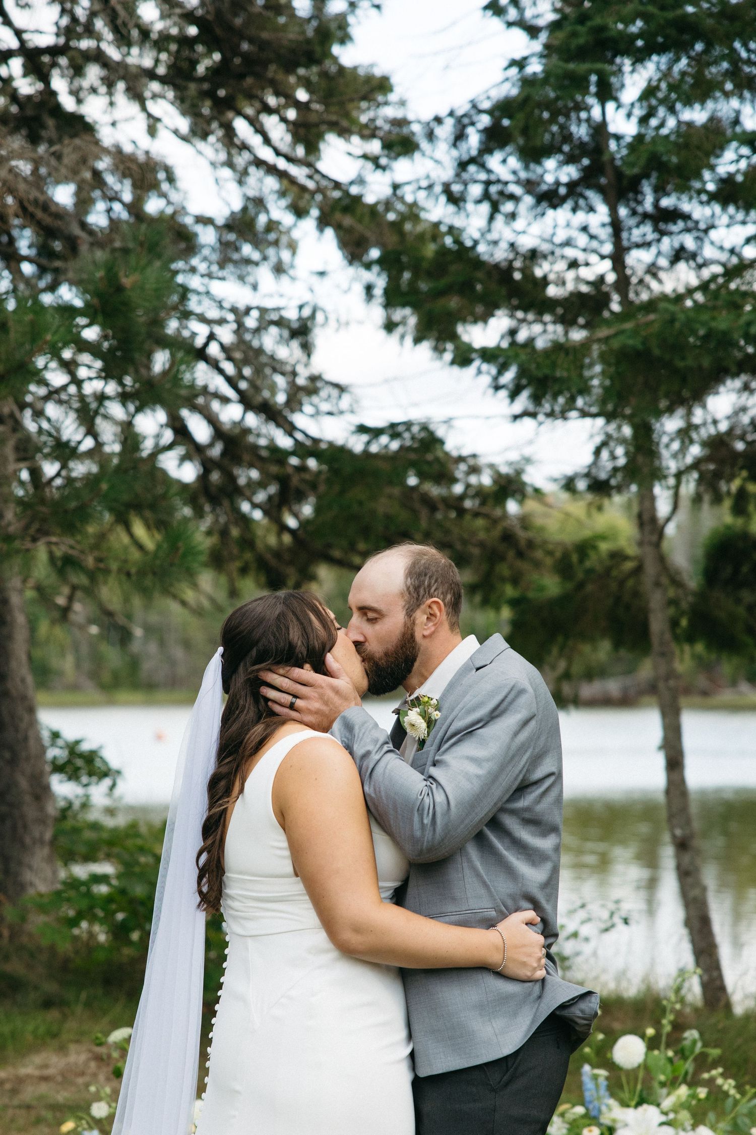 Romantic wedding moment captured by the lake with evergreen trees in background.