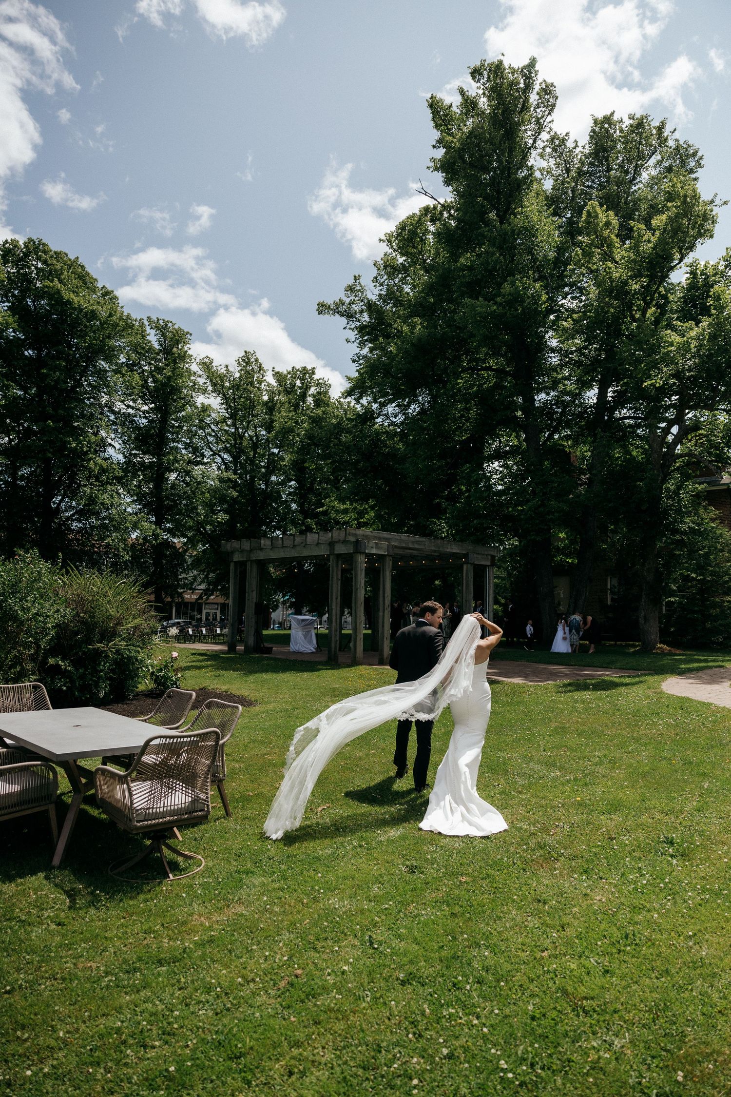 A flowing white wedding veil catches the breeze in a sunny garden setting with outdoor furniture.