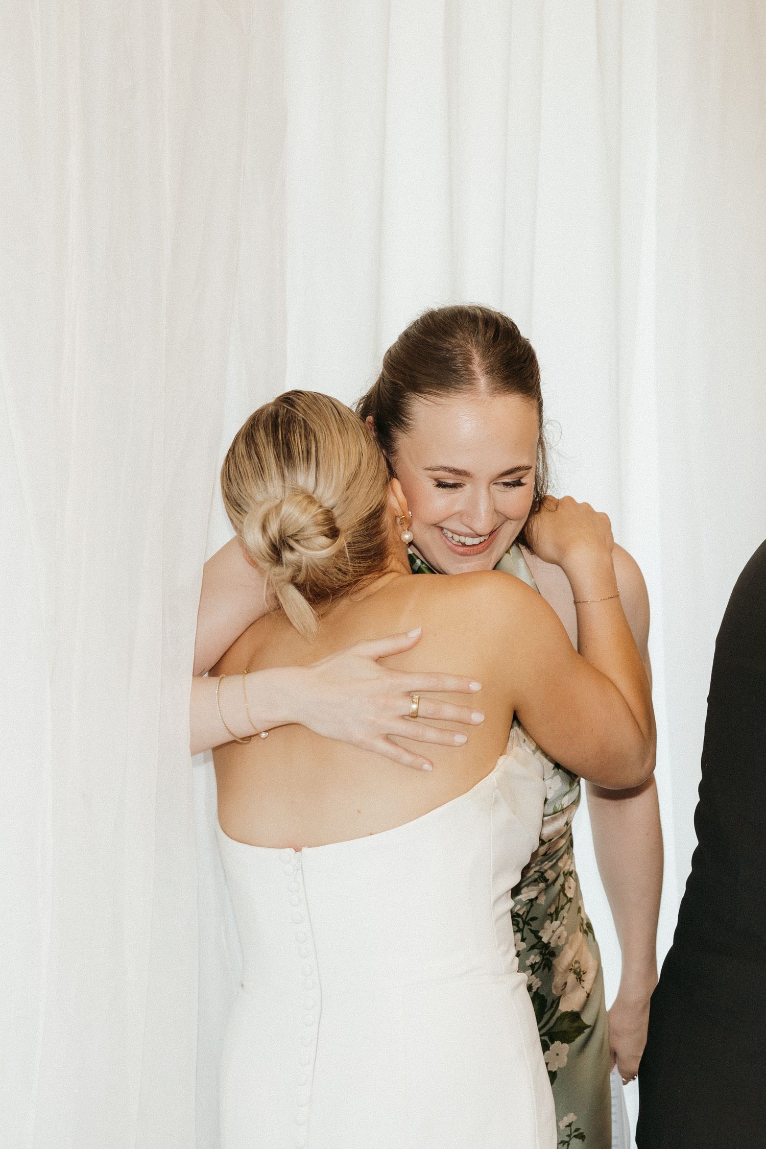 Two people share an emotional embrace in front of white curtains at a wedding celebration.