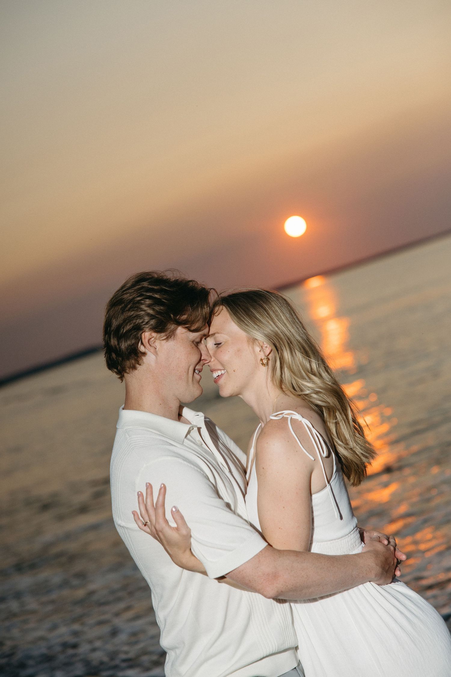 Couple embracing and smiling at each other on the beach during a stunning orange sunset.