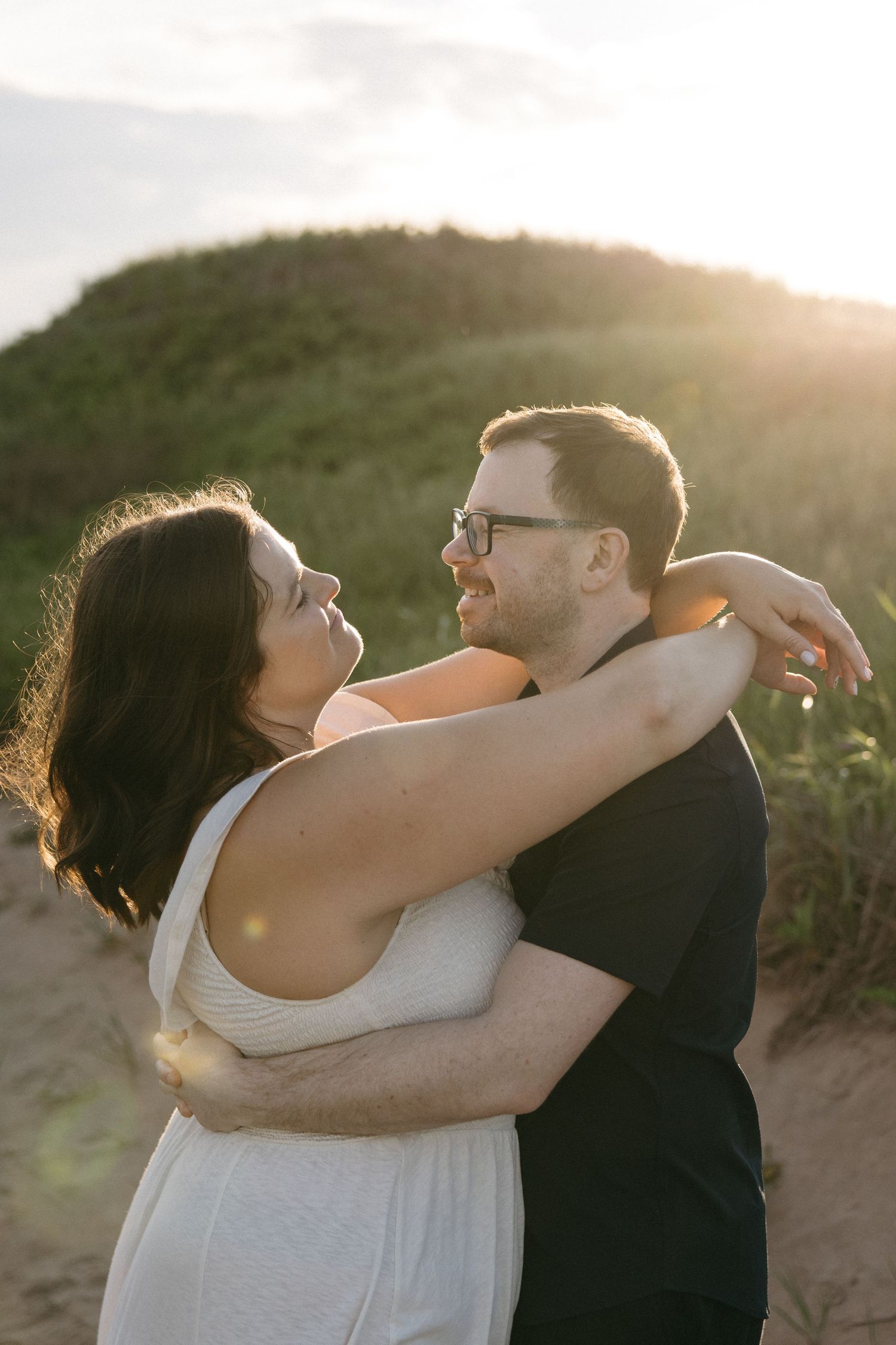 Couple embracing at golden hour on a sandy beach, woman in white dress smiling up at man in black shirt wearing glasses.