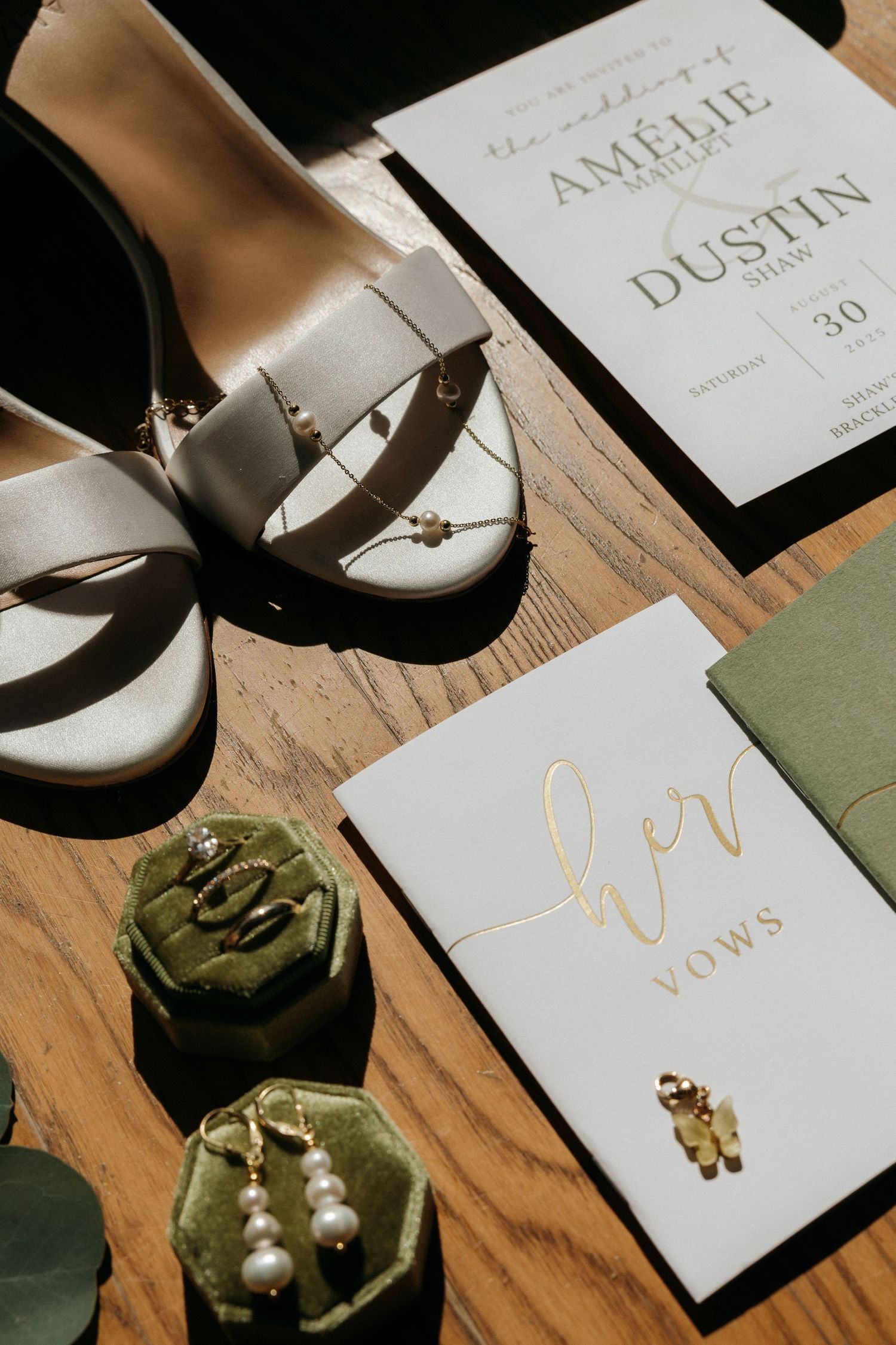 Wedding flat lay with white heels, green velvet ring boxes, pearl earrings, vow book, and elegant invitation suite on wood.