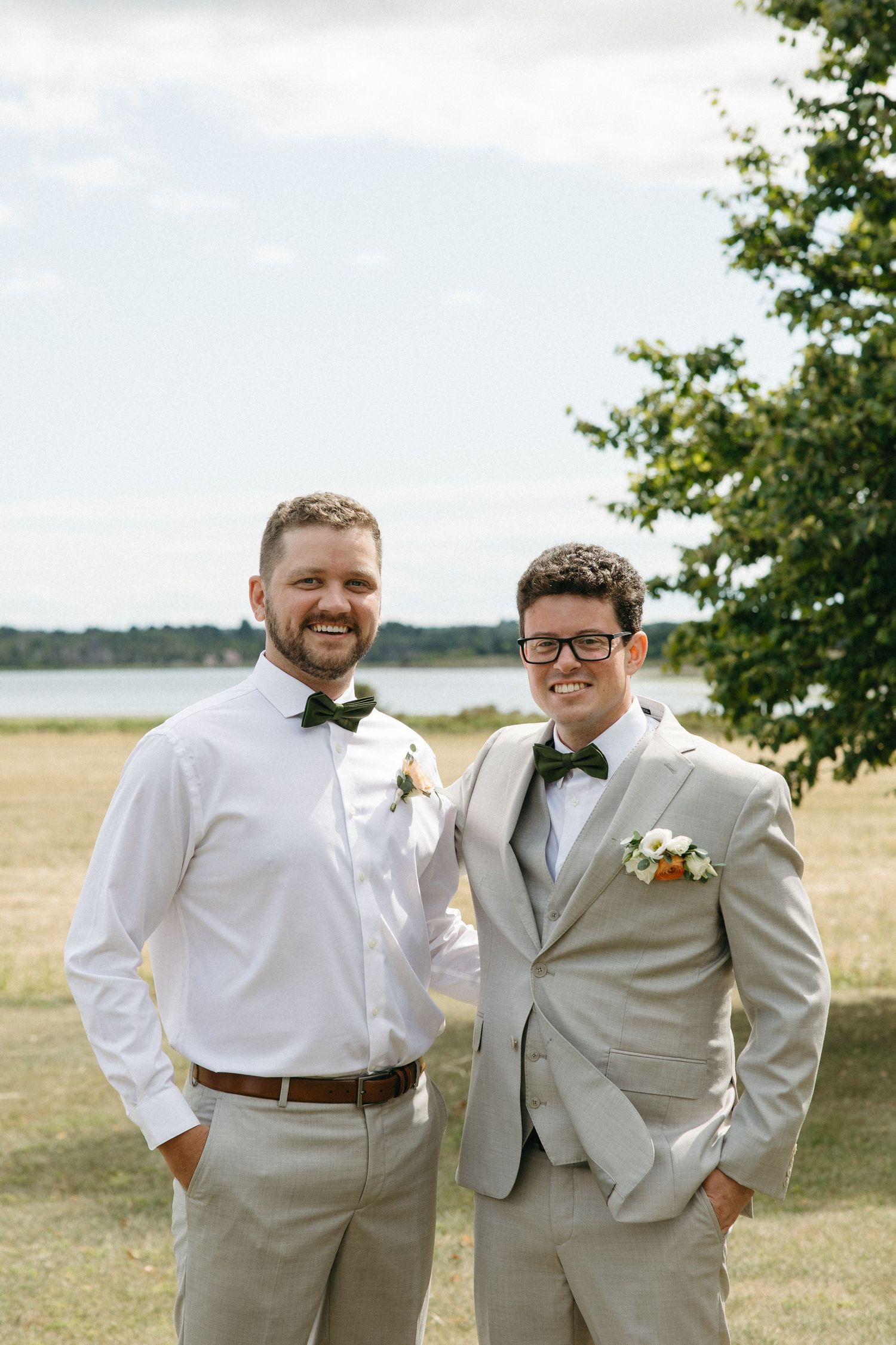 Two groomsmen in matching gray suits and bow ties posing together near a lake on a wedding day.