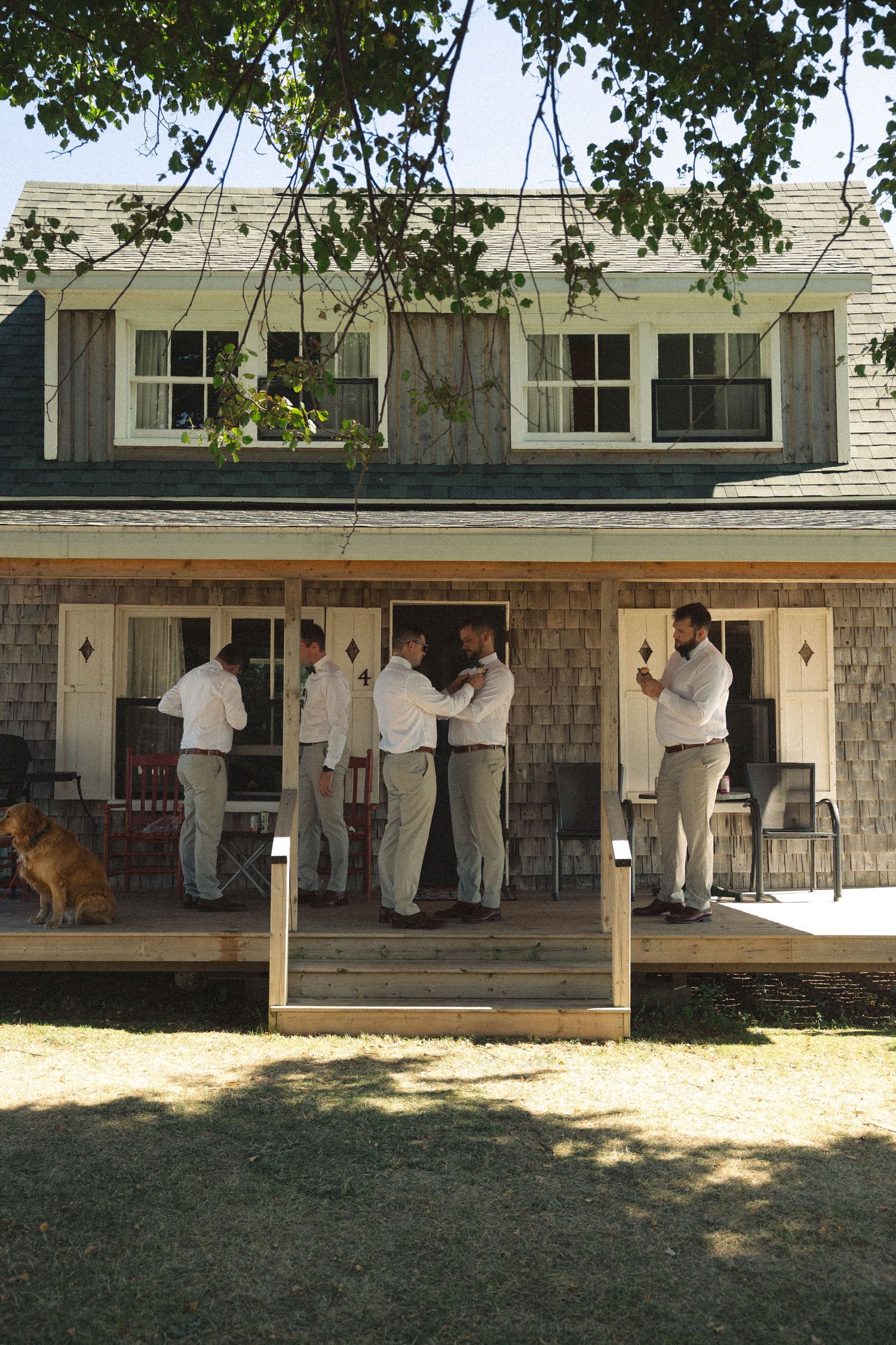 Groomsmen in white shirts and gray pants prepare on a rustic wooden porch of a shingle-style home.