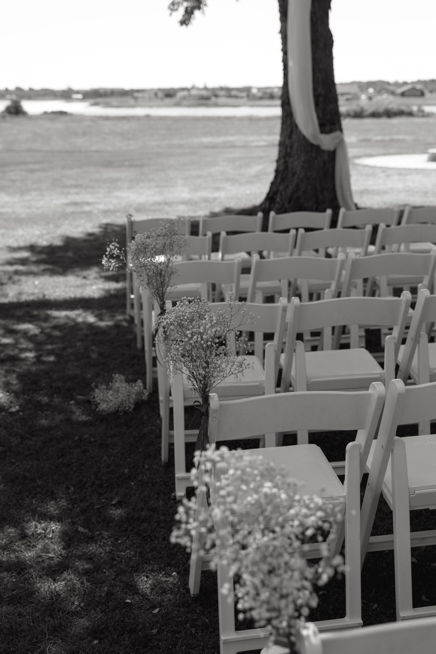 Rows of white folding chairs set up outdoors for a wedding ceremony near a tree.