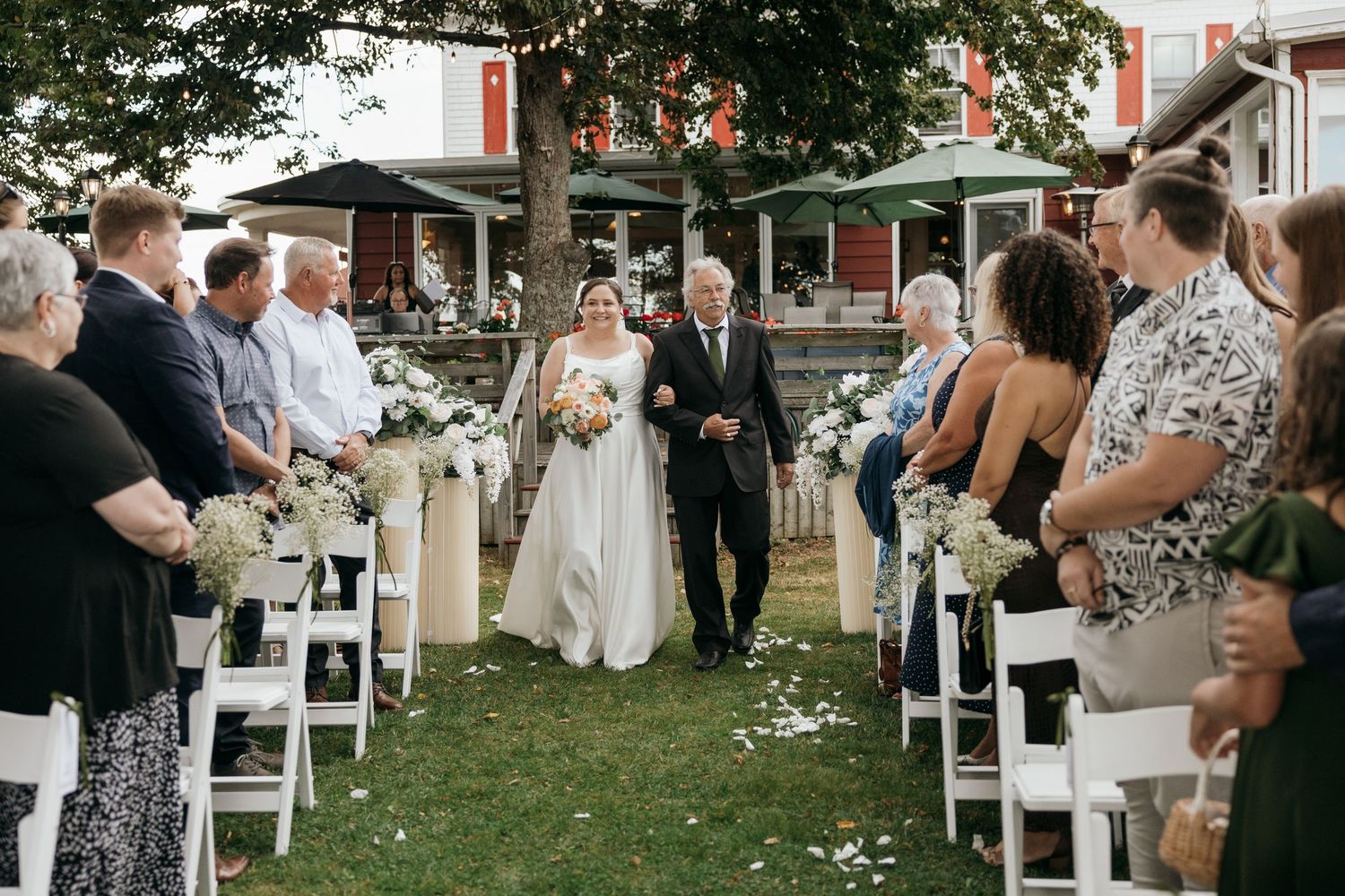 Bride walks down outdoor aisle with escort past seated guests in white chairs at garden wedding ceremony.