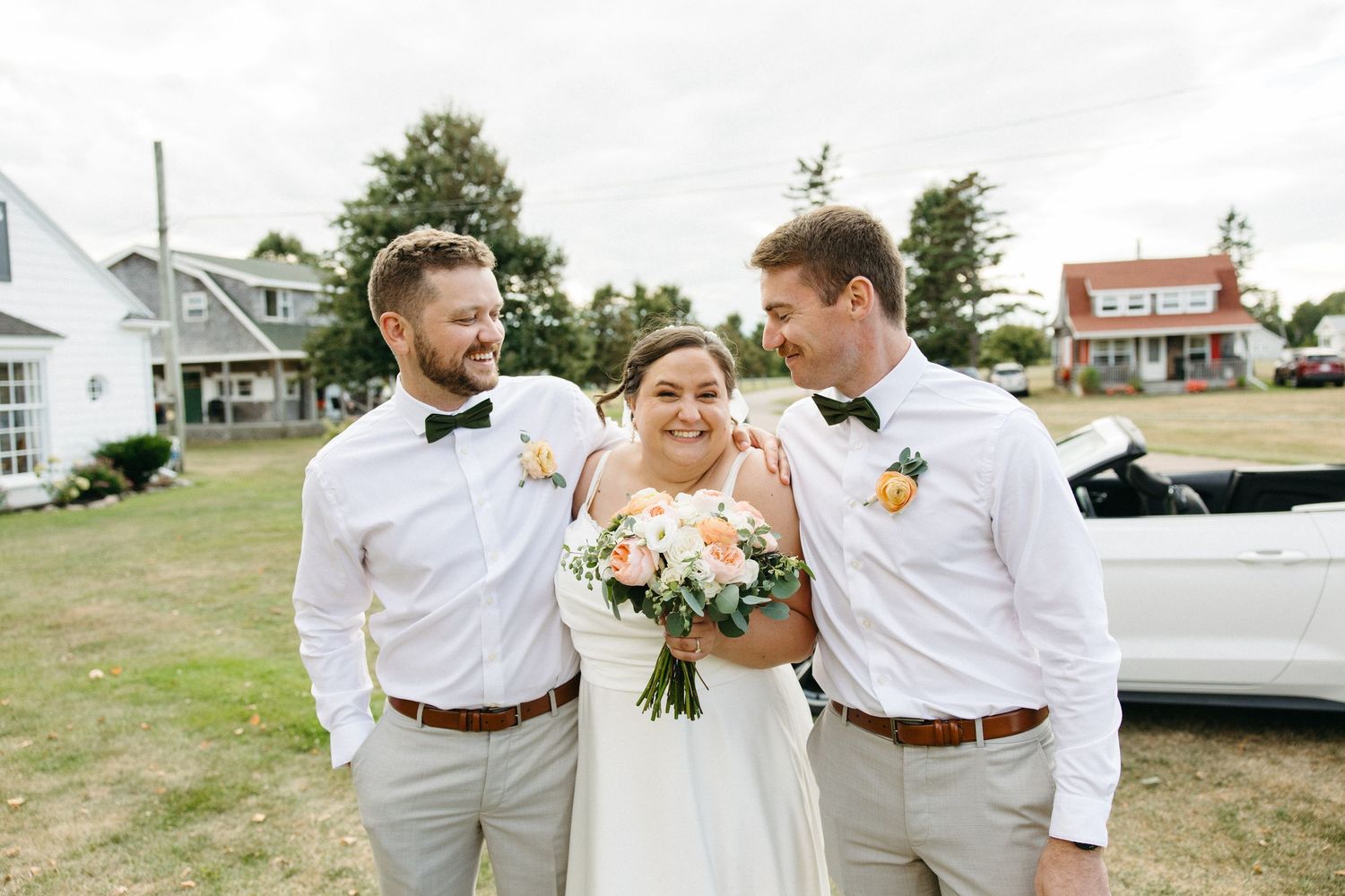 Smiling bride with bouquet stands between two groomsmen in bow ties on a green lawn at outdoor wedding.