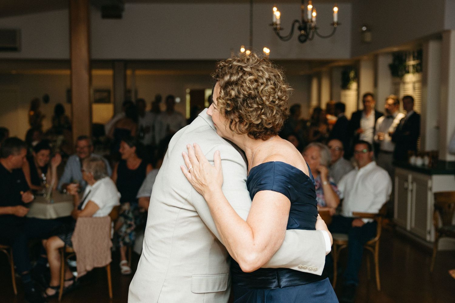 Groom shares tender mother-son dance at indoor wedding reception with guests watching in background.