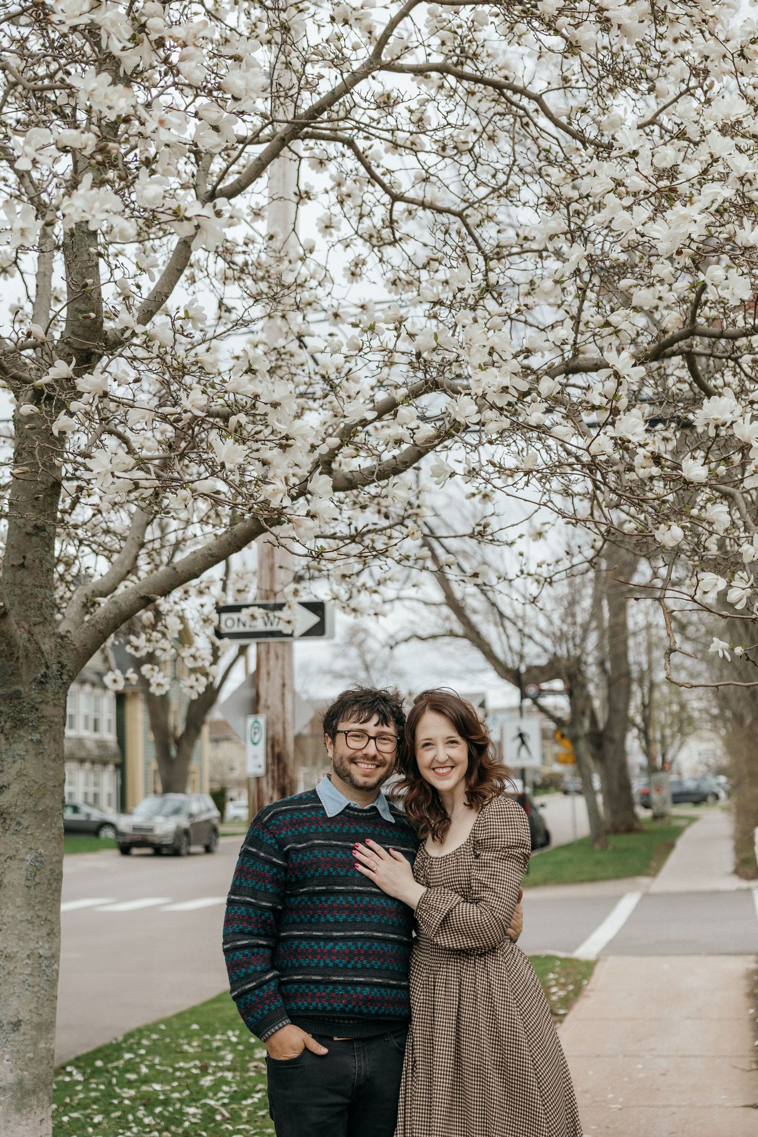 A smiling couple embraces under blooming white magnolia trees on a charming city sidewalk in spring.